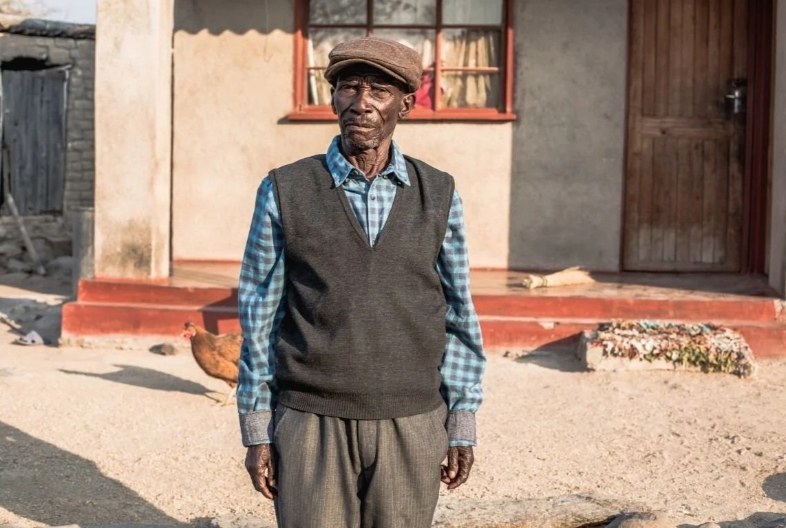 Fred outside his simple home. He is wearing a flat cap and there is a chicken in the background