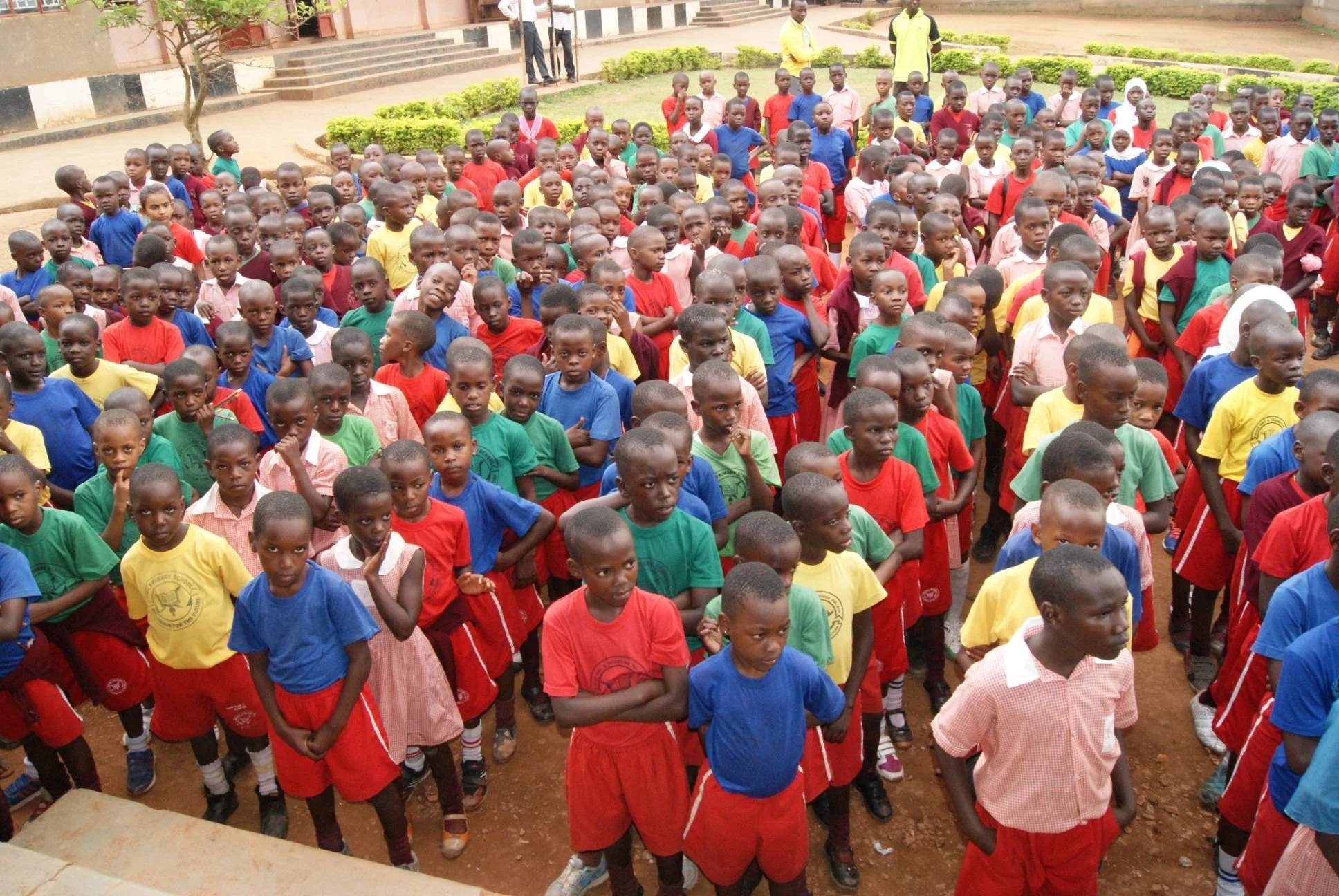 A large group of school children gathered outdoors, wearing colorful uniforms, standing on a dirt ground in front of a building with steps and greenery.