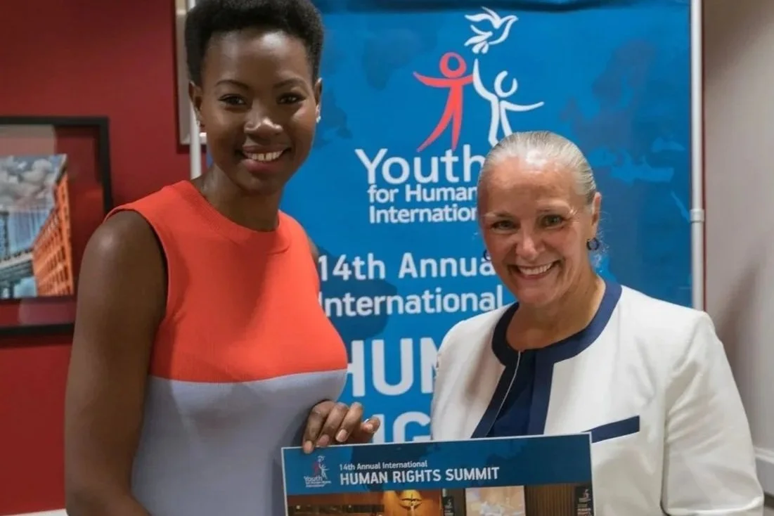 Two women smiling at the camera during the 14th Annual International Human Rights Summit, with a blue Youth for Human International banner in the background.