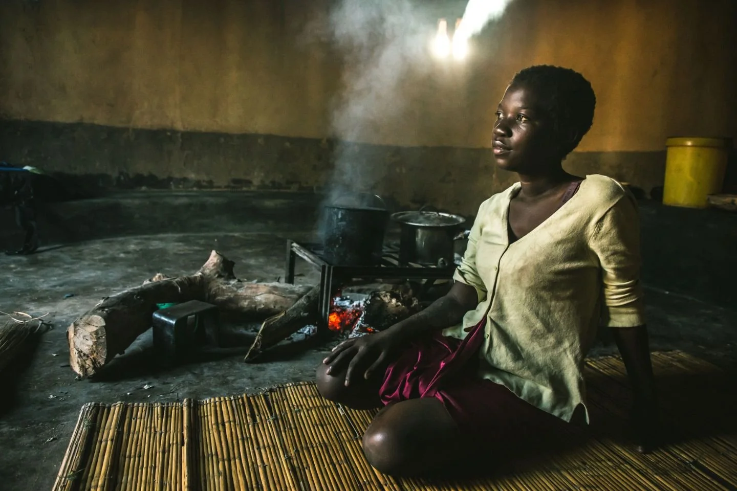 A woman sitting on a mat near an open fire inside a simple, rustic room. The woman is looking to her right. The room has a yellow wall, and some pots on the stove, with smoke rising from the fire.