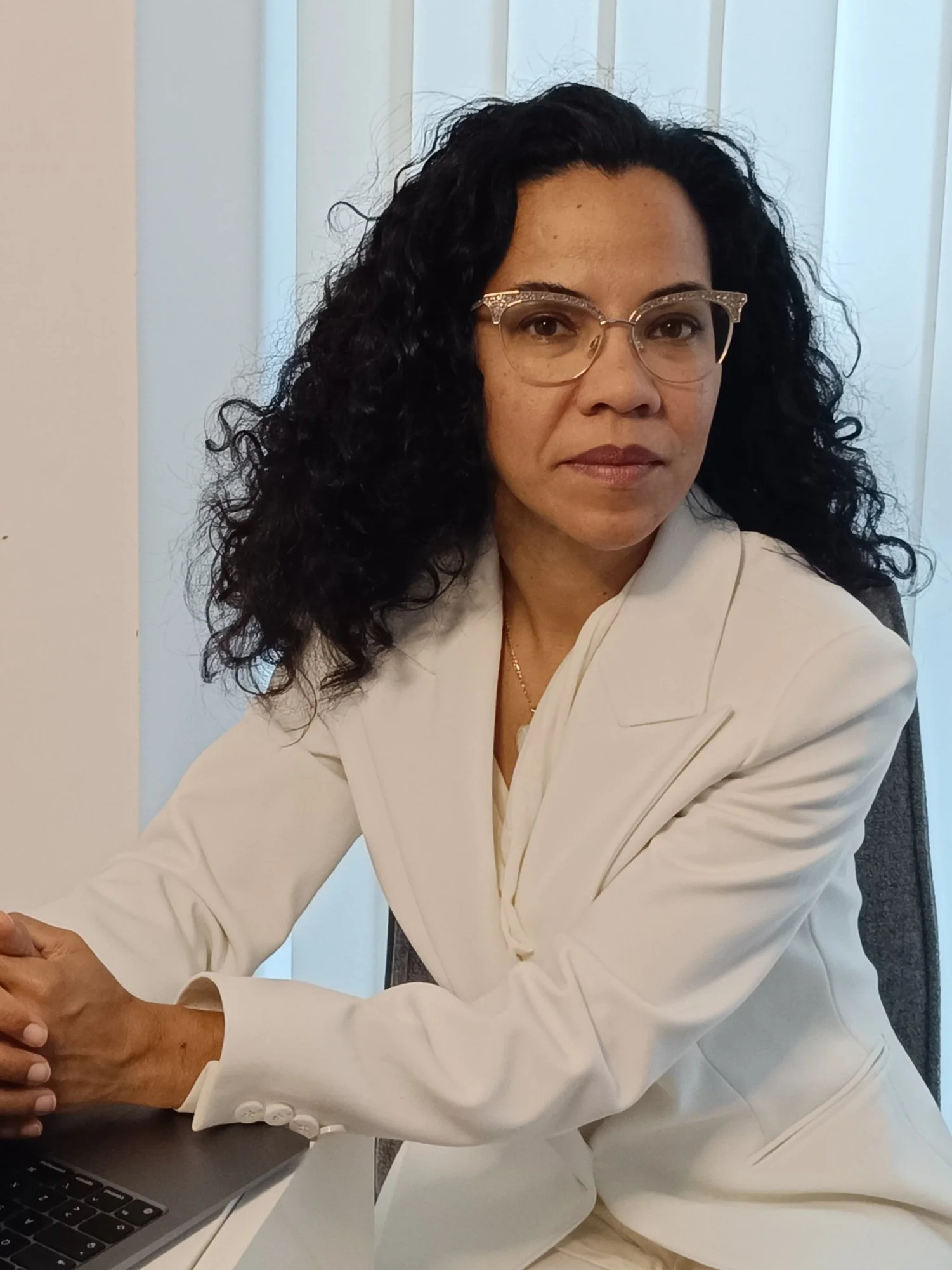 Mujer con cabello rizado y gafas, vestida con traje blanco, sentada en una oficina frente a una computadora portátil.