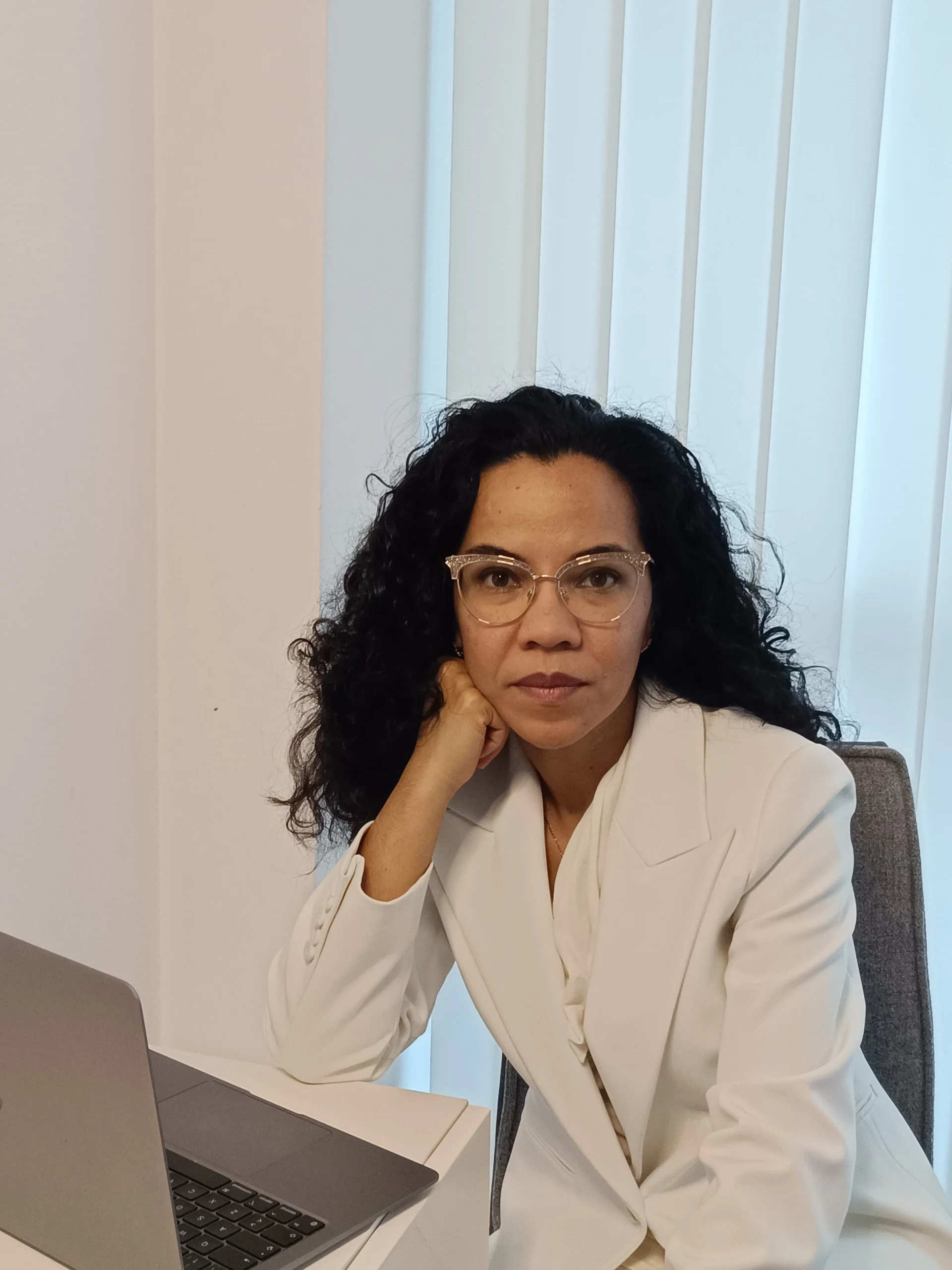 Mujer con cabello rizado oscuro, gafas transparentes, ropa blanca y expresión pensativa, sentada en una oficina con laptop y persianas blancas detrás.