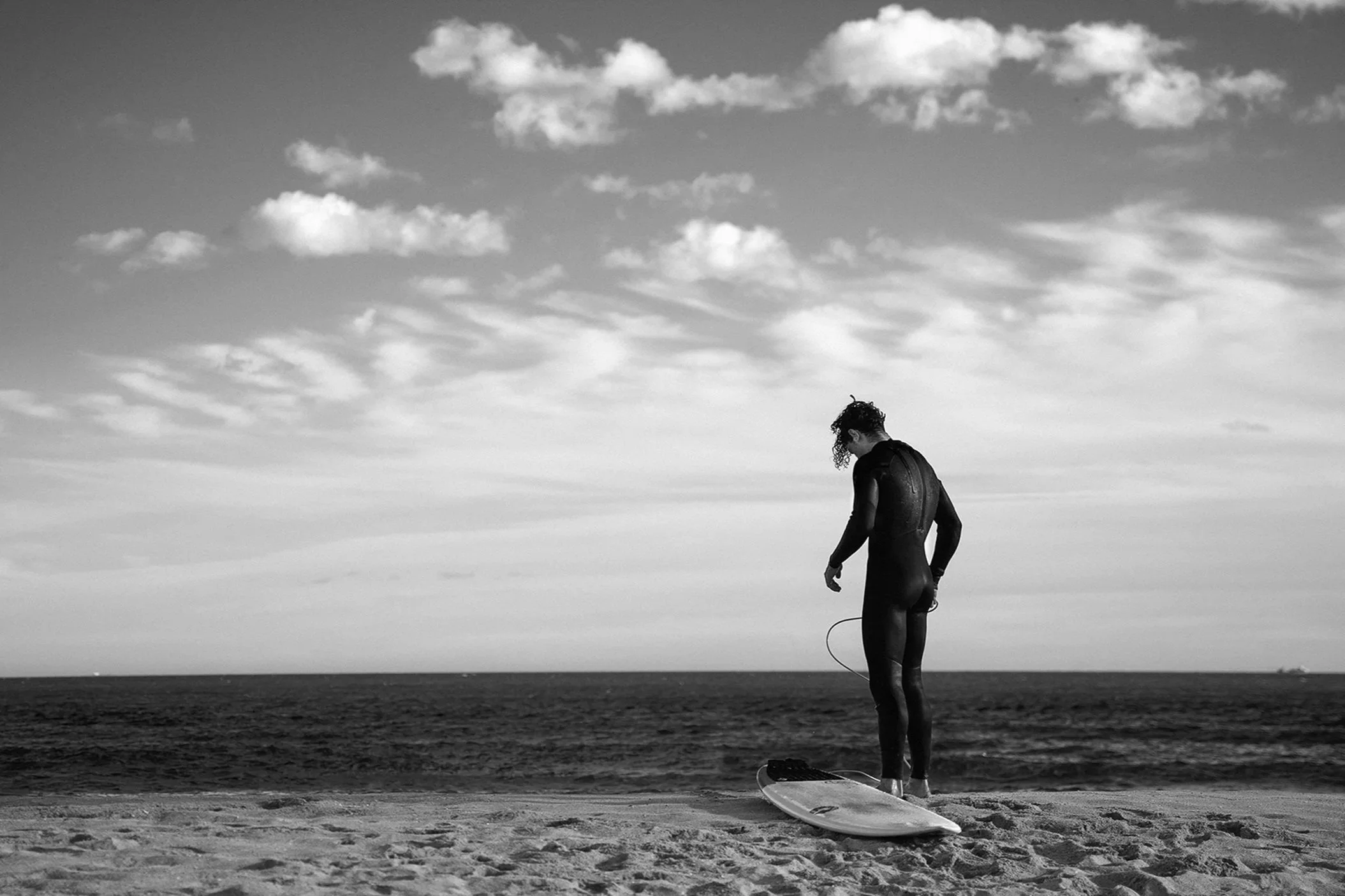 A woman in a wetsuit standing on a surfboard on the beach, looking down, with the ocean and cloudy sky in the background.