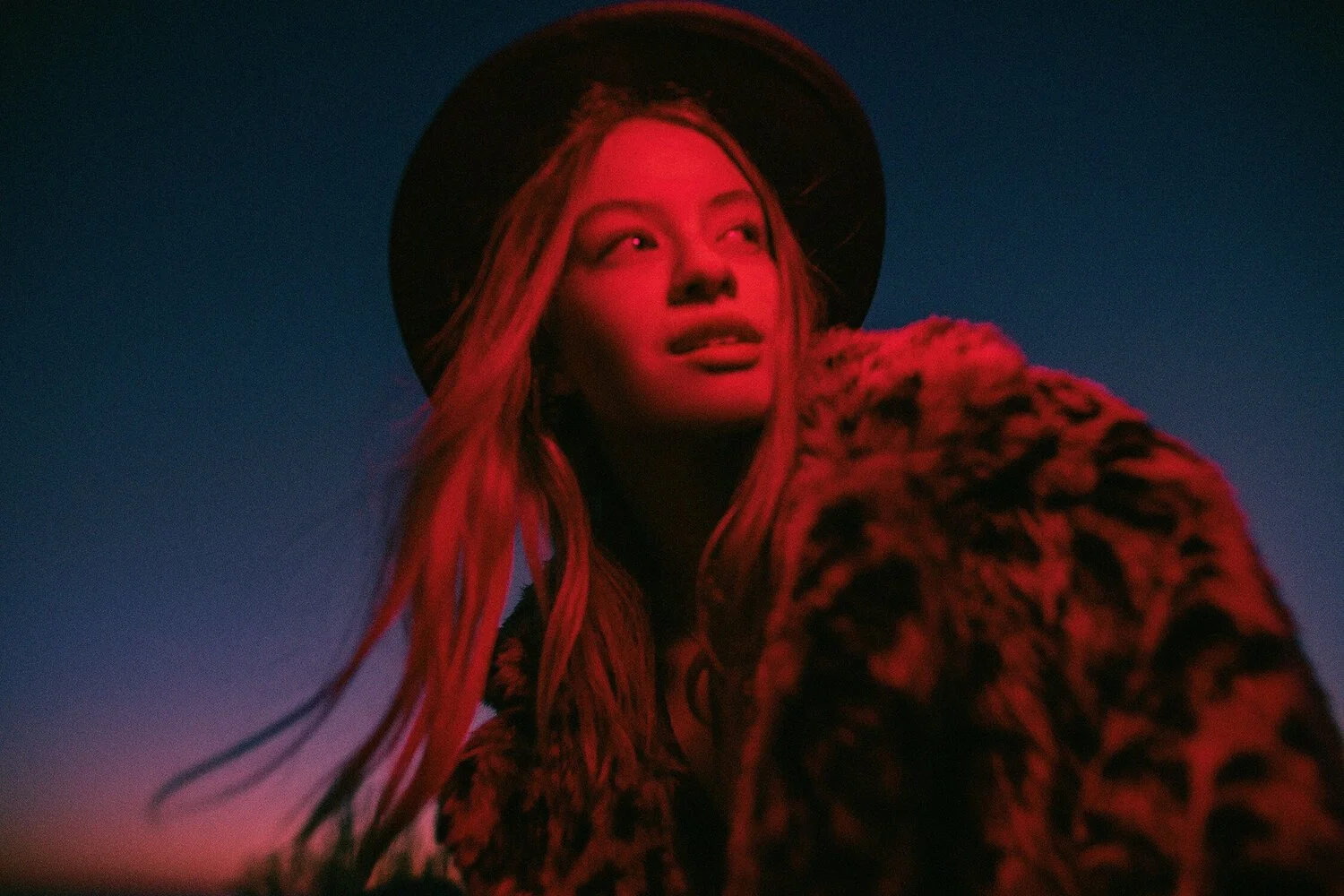 A woman with long red hair wearing a black hat and leopard print coat, looking up with a slight smile against a dark blue and pink gradient sky. Fashion photographer in Barcelona.
