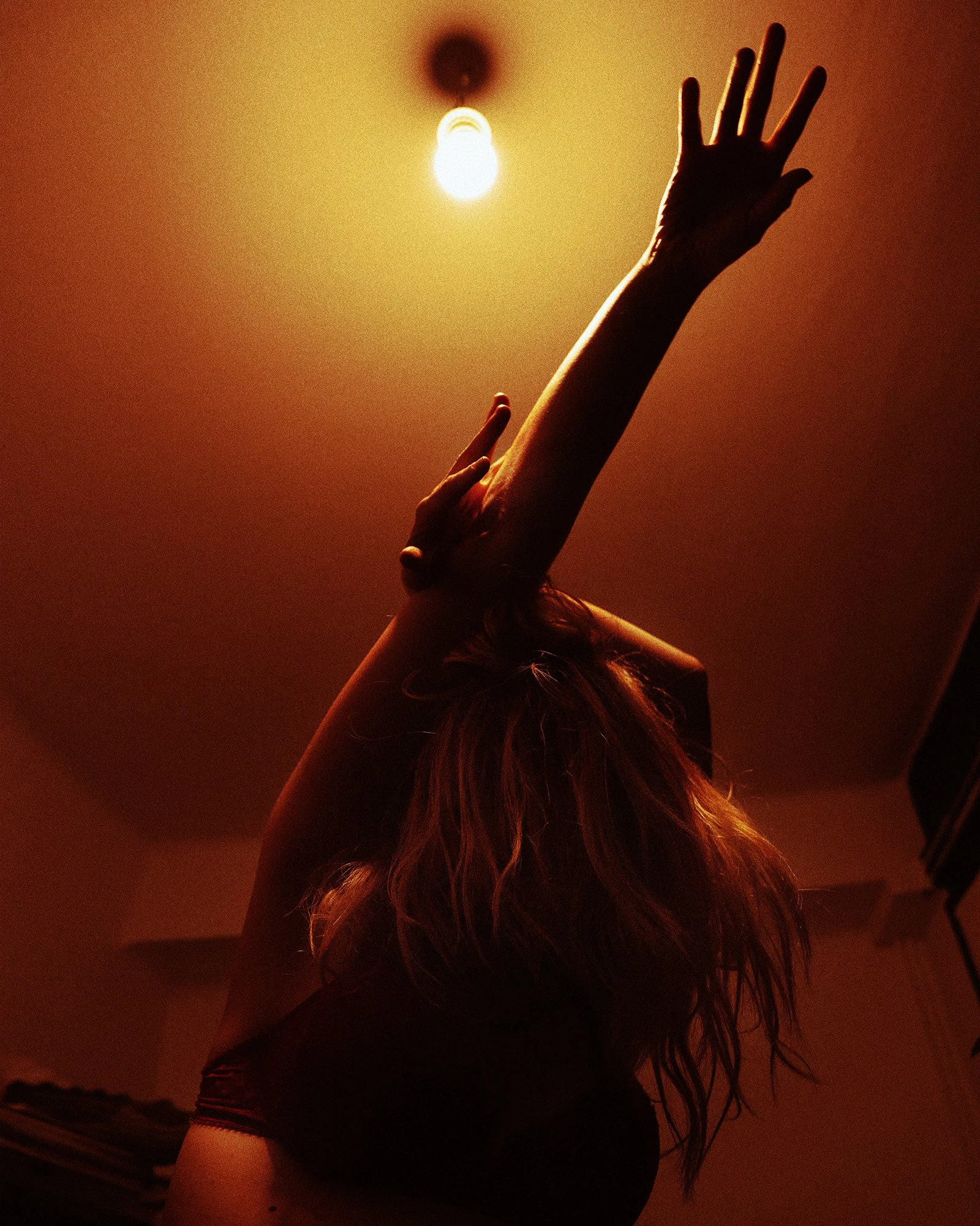 A person with long hair reaching toward a bright light bulb hanging from the ceiling.