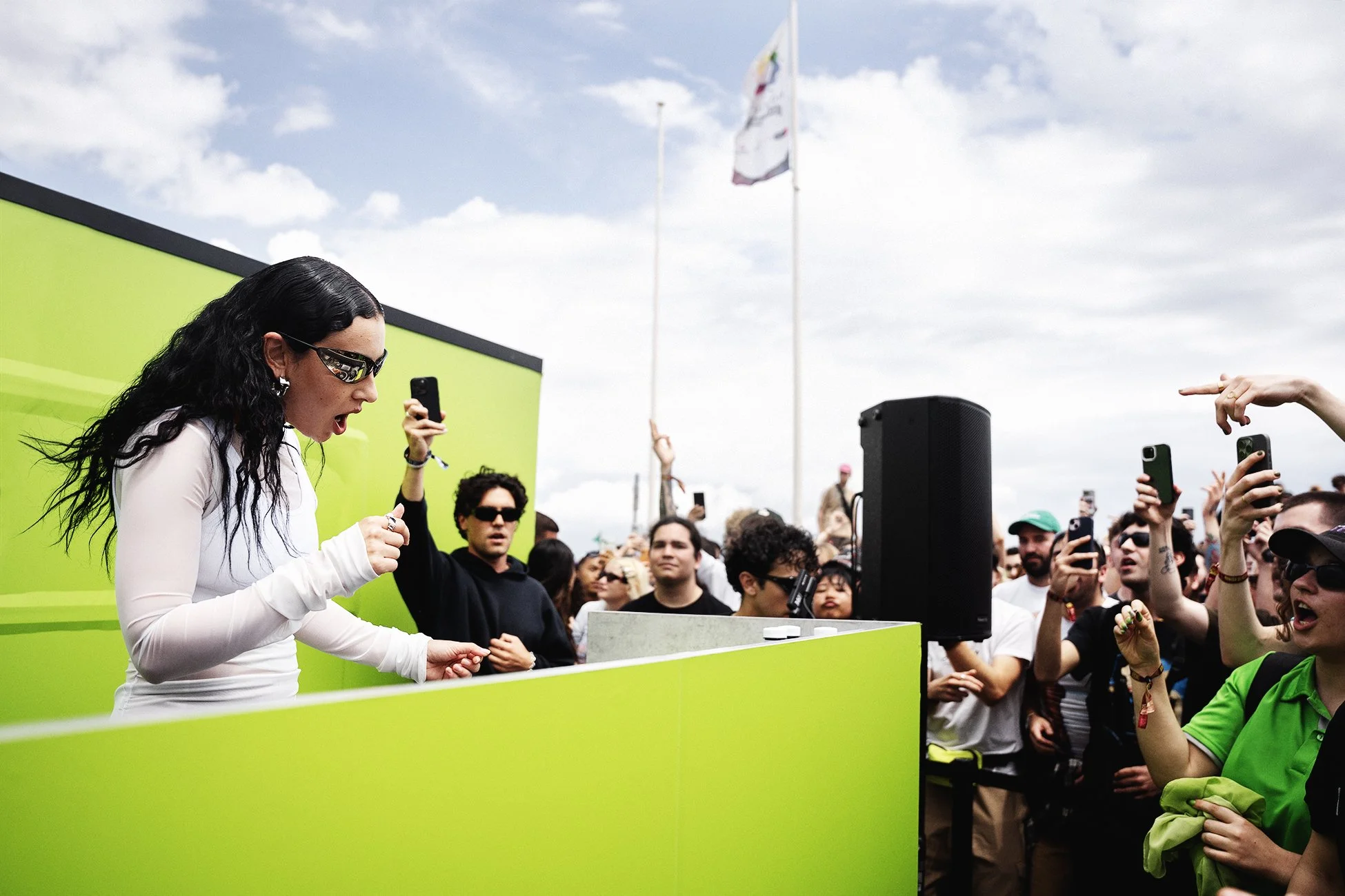 A woman with black curly hair, wearing a white long-sleeve shirt and sunglasses, appears to be speaking or singing passionately at an outdoor event, with a crowd of people. The crowd is diverse. Event photographer in Barcelona.