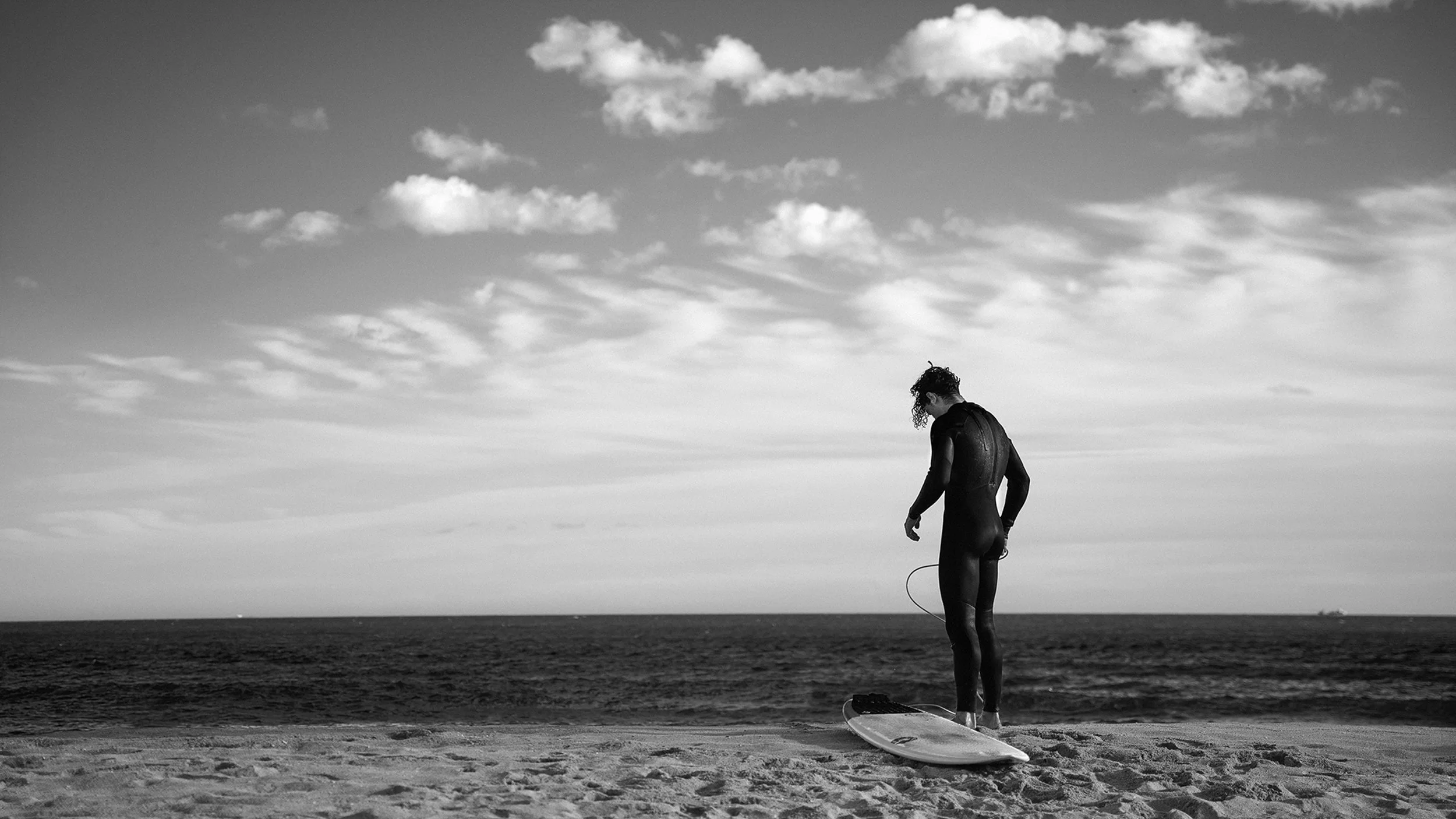 A woman in a wetsuit standing on a surfboard on the beach, looking down, with the ocean and cloudy sky in the background.