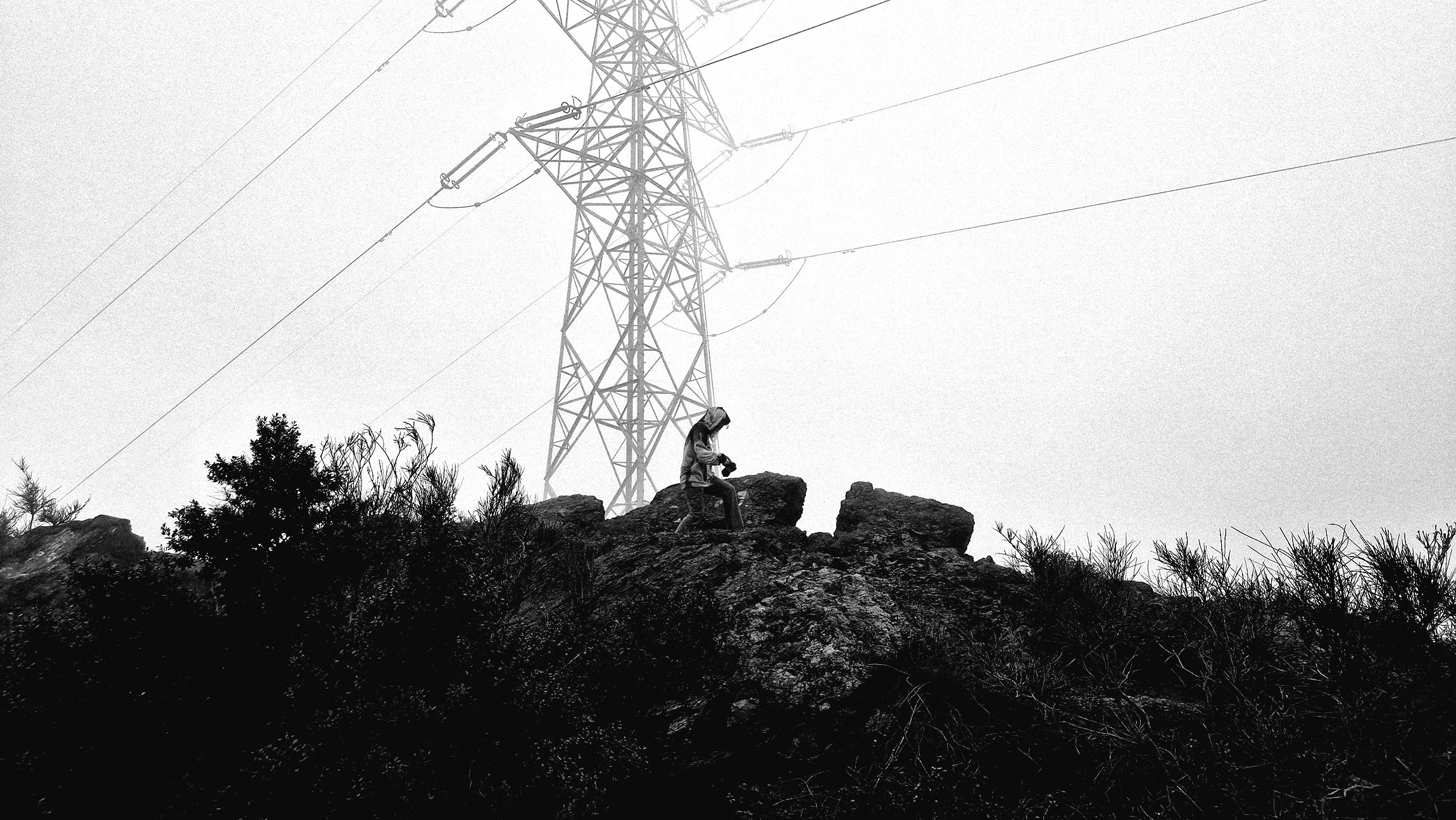 A person sitting on rocks with a large electrical transmission tower in the background, overcast sky.