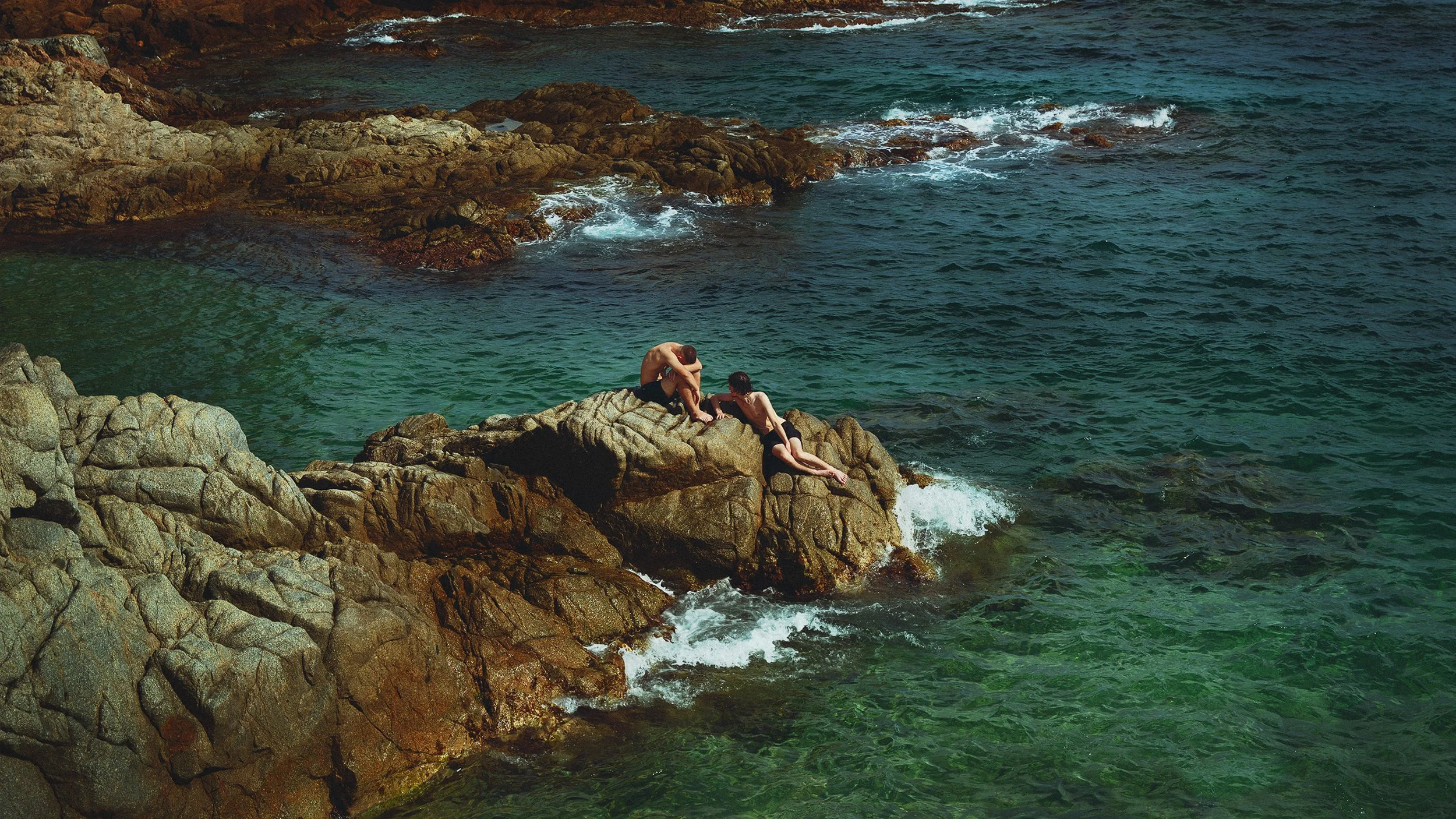 Two children are sitting on large rocks by the ocean, with one child lying down and the other leaning over, near the water's edge.