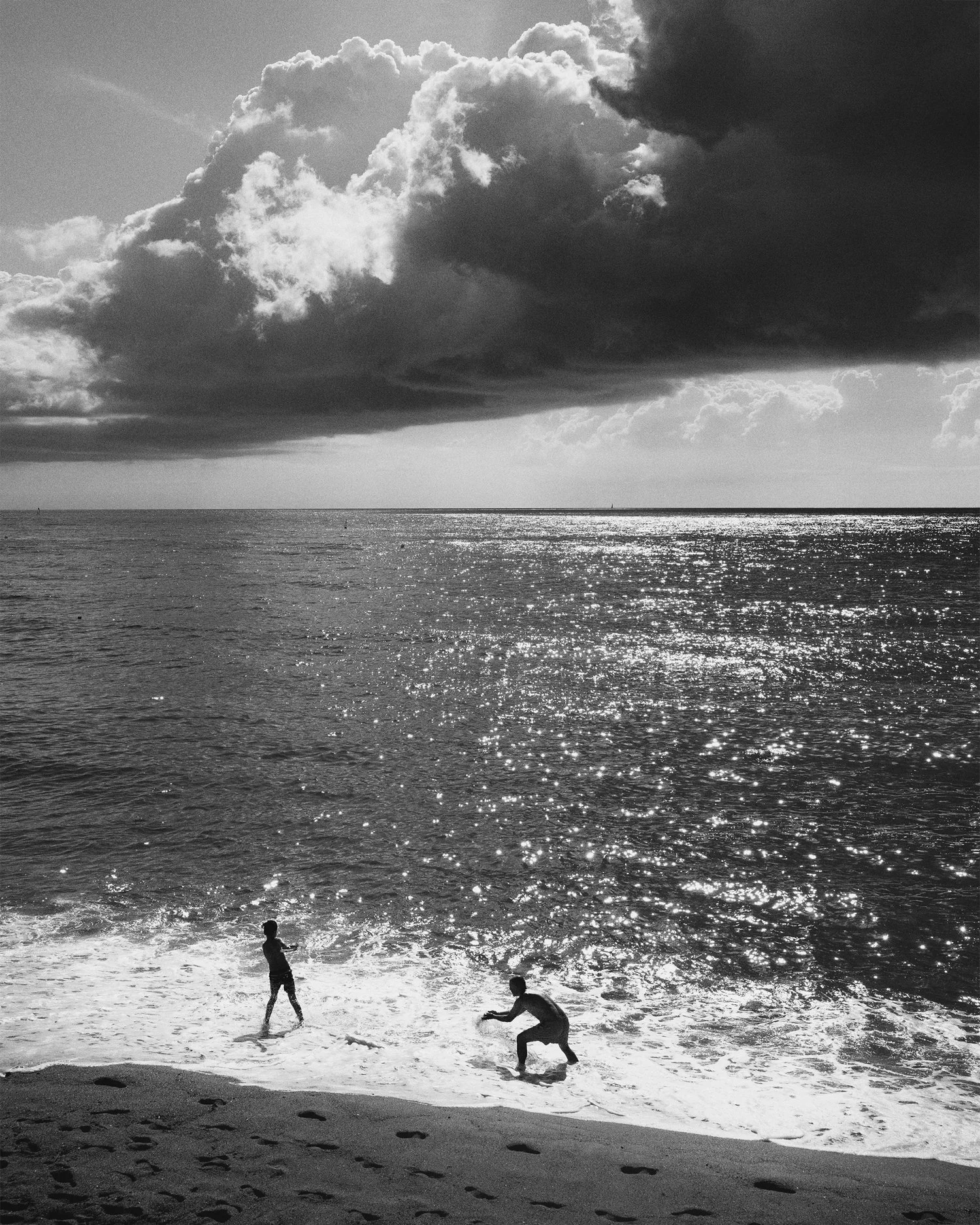 Black and white photo of a beach with some children playing near the shoreline, and a large cloud covering part of the sky above the water, reflecting sunlight.
