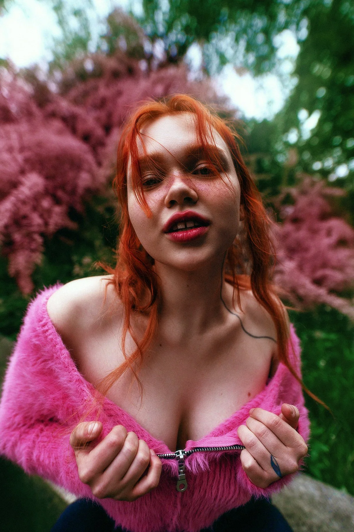 A woman with red hair wearing a pink fuzzy jacket, holding the zipper with both hands, outdoors with pink flowering trees in the background. Fashion photographer in Barcelona.
