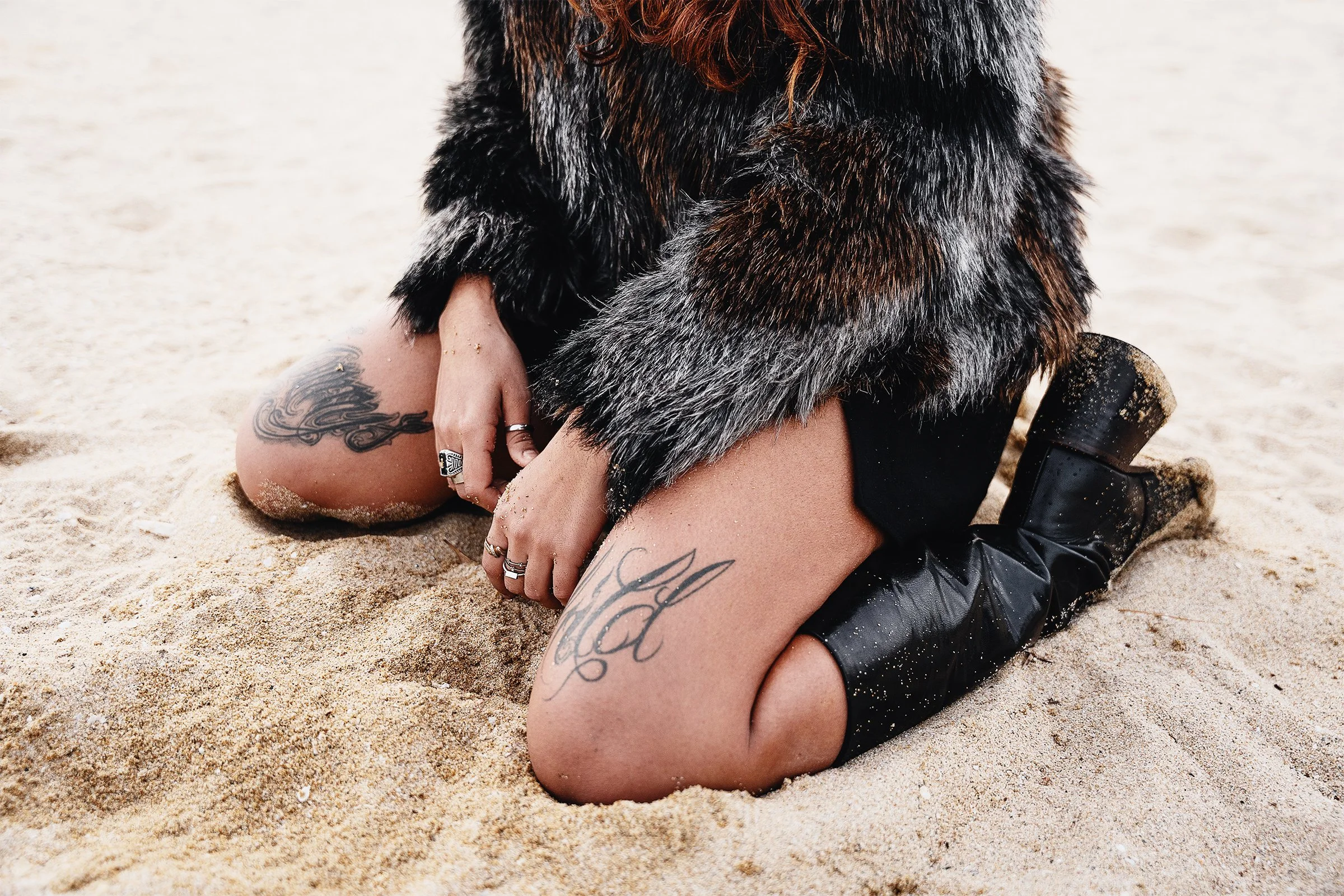 Person kneeling on sandy beach, wearing black thigh-high boots, black shorts, a fur coat, and displaying tattoos on thighs and hand. Fashion photographer in Barcelona.