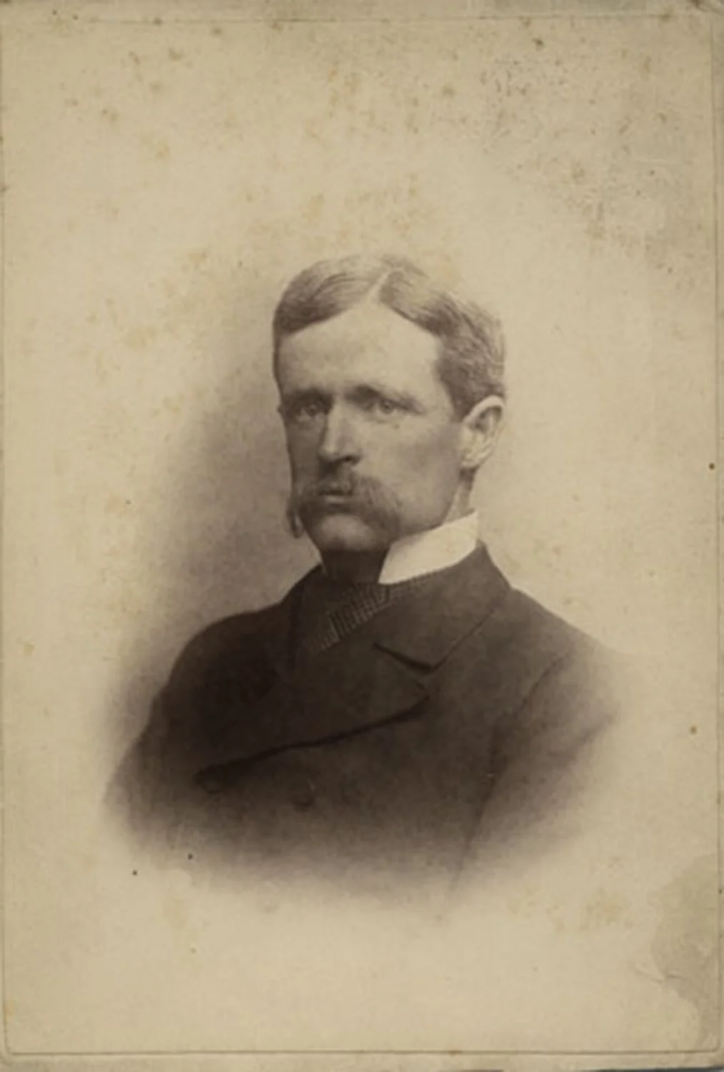 A black-and-white photo of a young man in a dress shirt and tie standing next to a large vintage camera or early film projector.