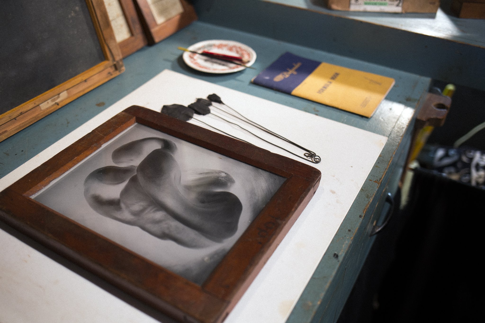 A glass plate shows a human ear pressing against it, figuratively creating an impression. The plate is on a white work surface, with brushes, a notebook, and a small plate in the background, indicating an art or craft workspace.