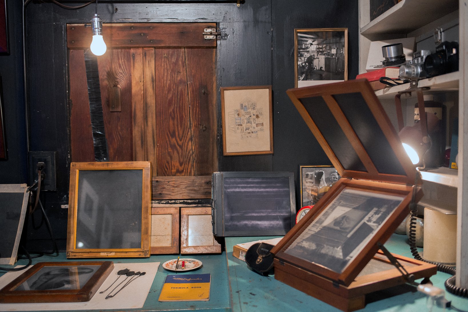 A cluttered workspace with various photographs, frames, and books, including a vintage camera on a shelf, a desk lamp, and a wooden door in the background.
