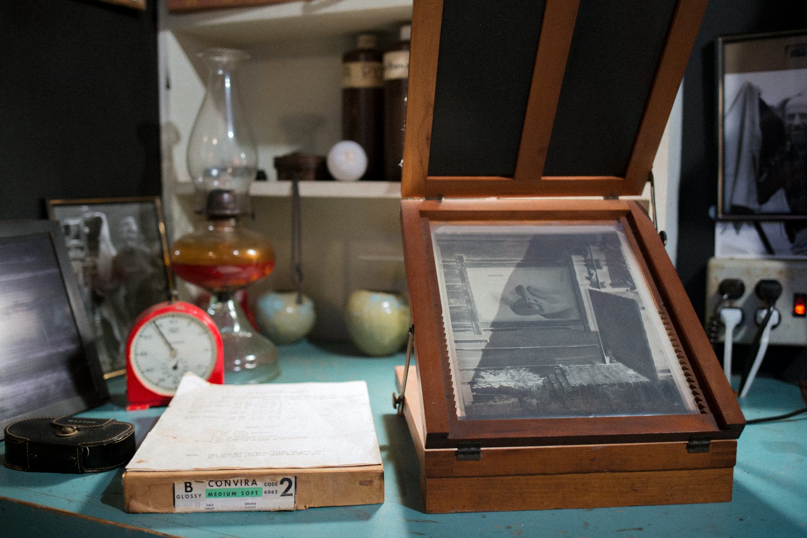 A table with vintage items including a wooden photo slide viewer, an open container with photographs, a red clock, a glass decanter, a black camera case, and a sheet of paper. In the background are shelves with jars and ceramics.