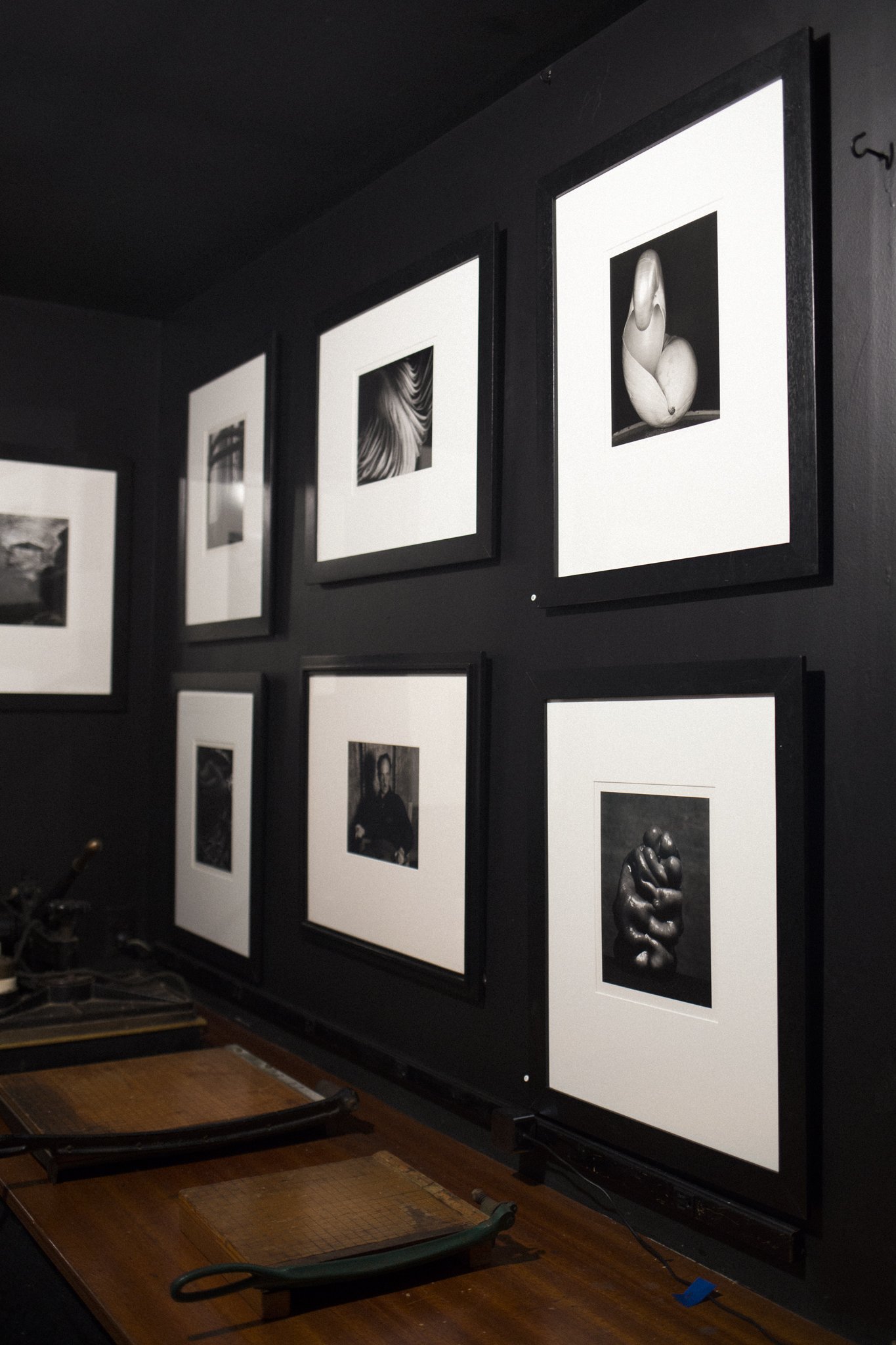 Black and white photographs displayed on a dark wall, featuring various artistic portraits and abstract images, with vintage tools or devices on a wooden surface below.