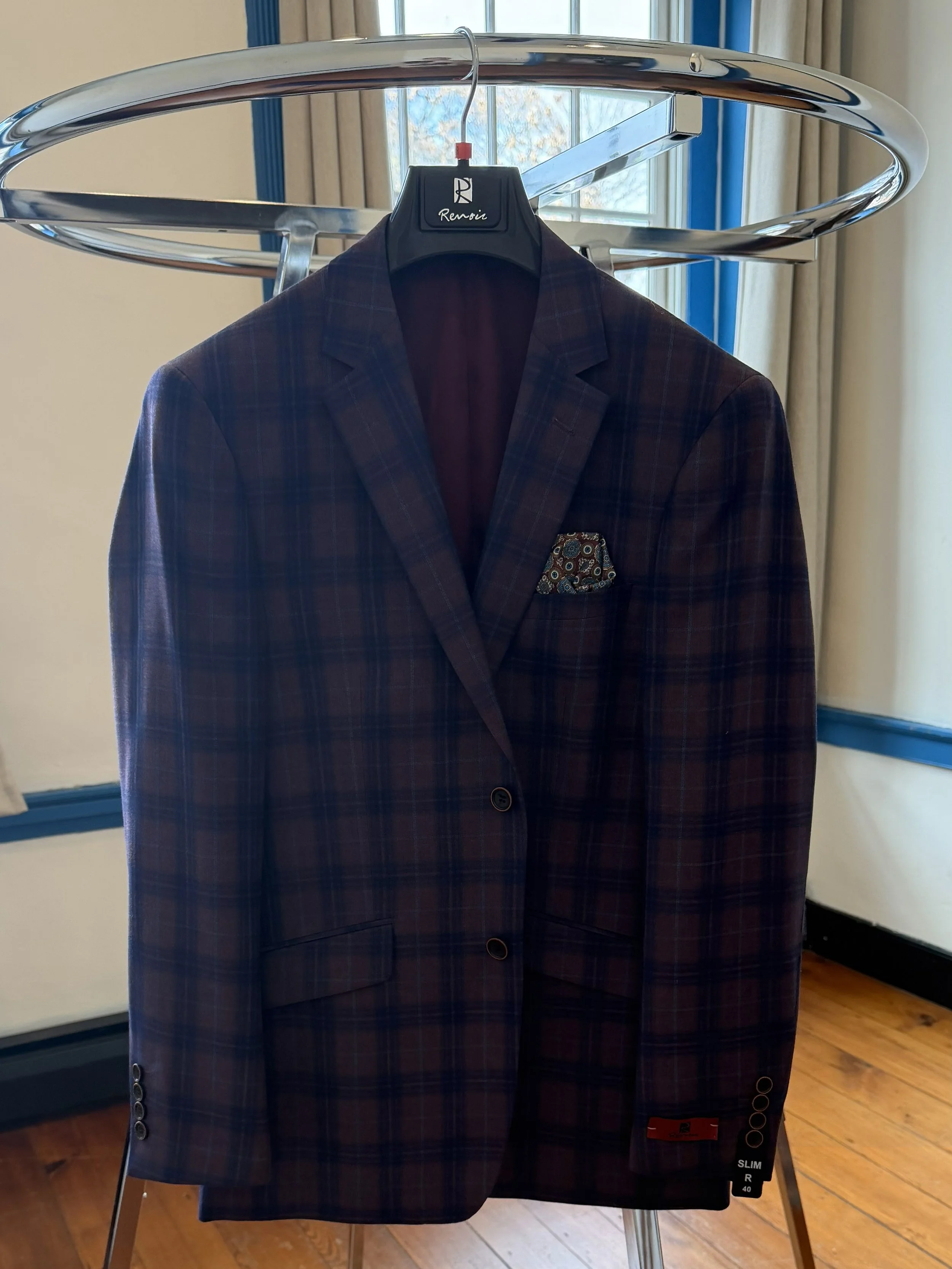 A plaid blazer jacket hanging on a black hanger on a chrome clothing rack, with a window and curtains in the background.