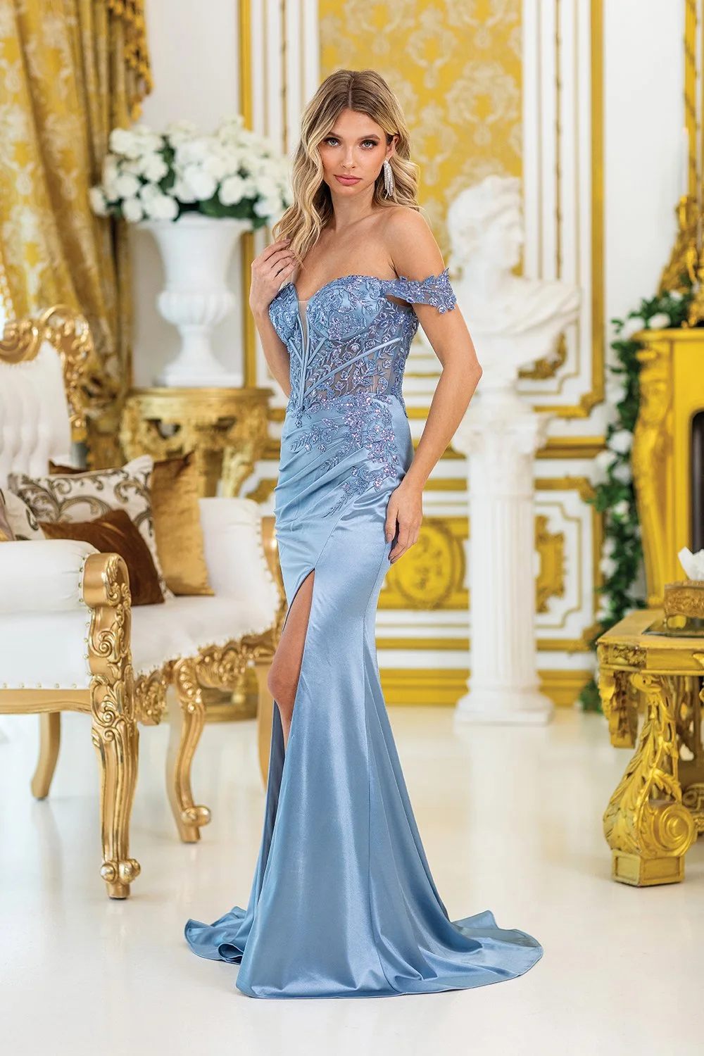 A woman in an elegant off-shoulder blue gown with lace and embroidery detail, posing in a luxurious, gold-accented room.