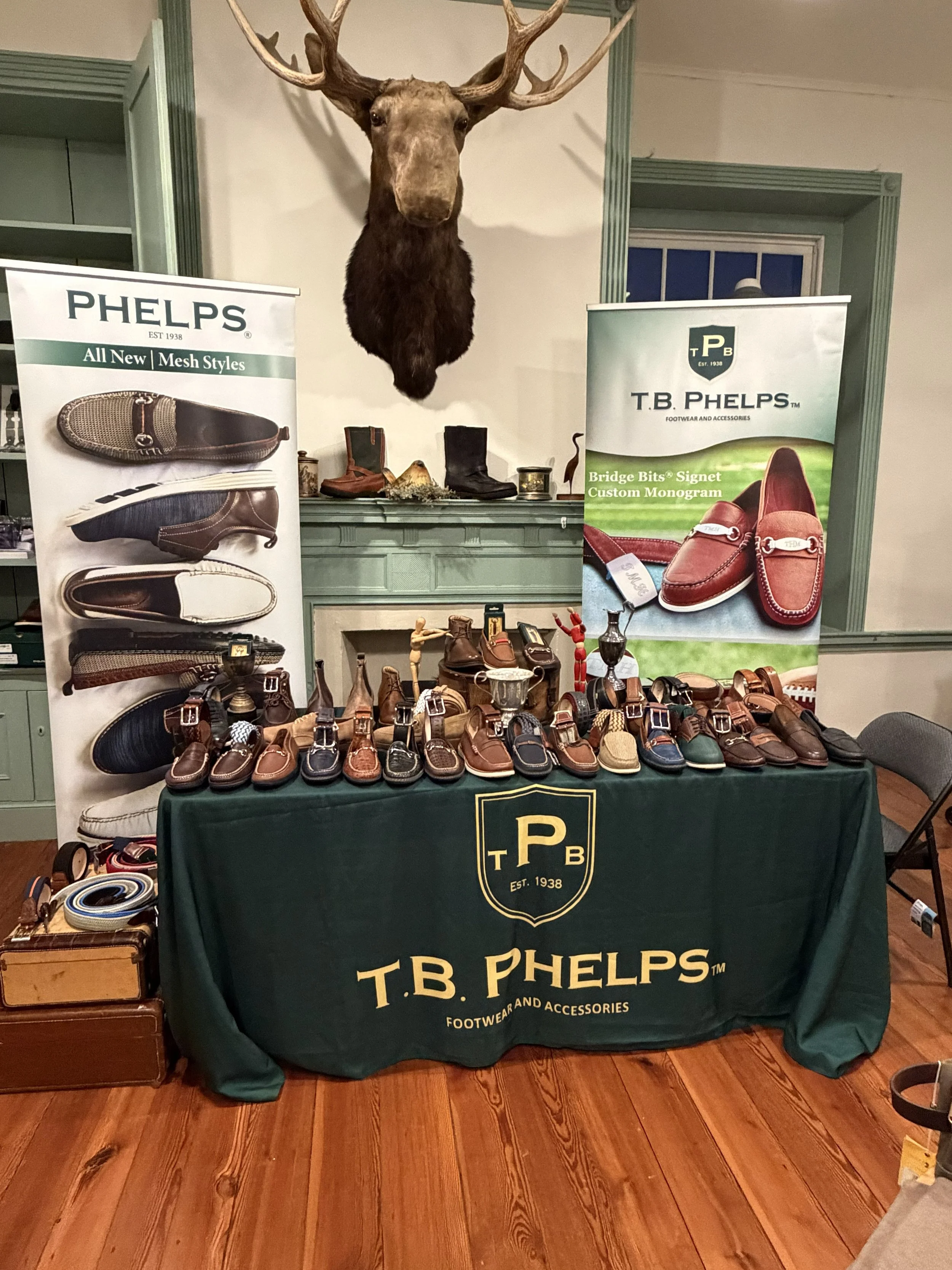 A display table featuring men's leather shoes and boots from T.B. Phelps, with banners advertising their styles and custom monogram options. A mounted moose head is on the wall behind the display, which includes a green tablecloth with the T.B. Phelps logo.