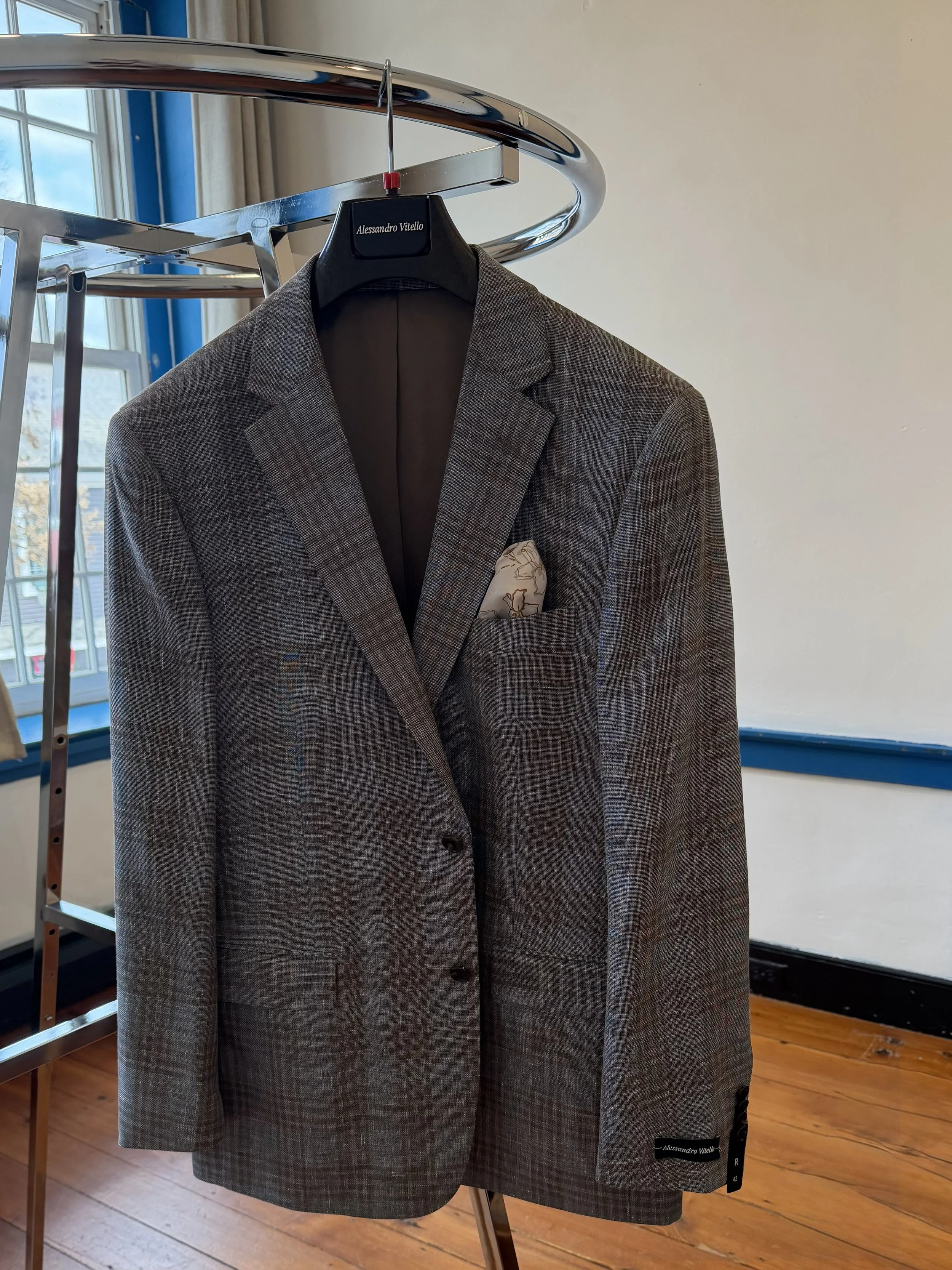Gray plaid men's blazer with a pocket square hanging on a metal clothing rack in a room with wooden floors and a window in the background.