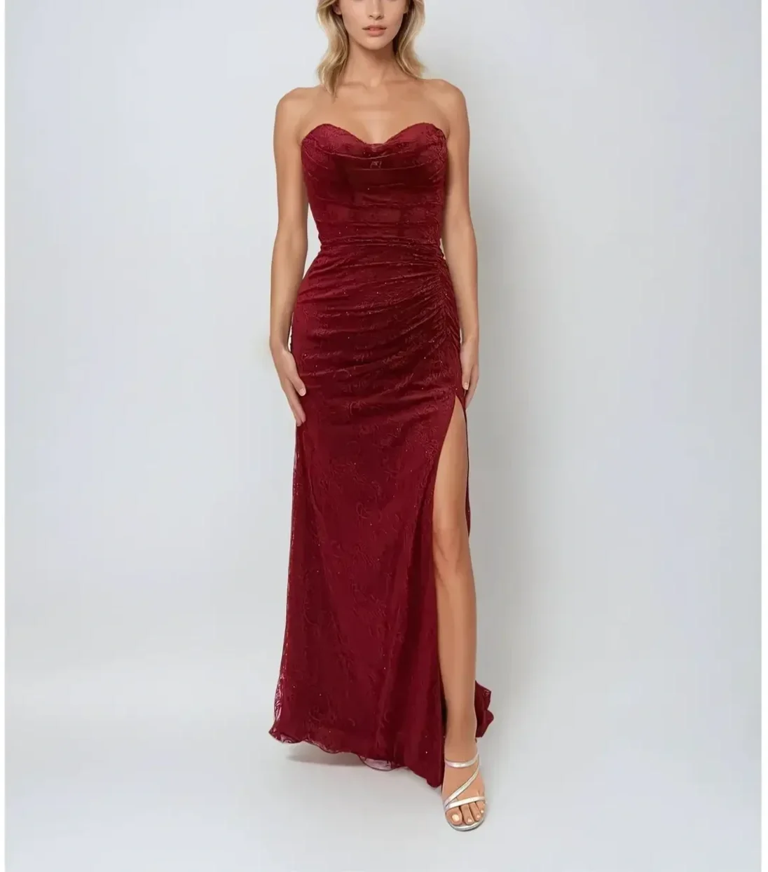 A woman wearing a strapless, deep red, ruched evening gown with a thigh-high slit, paired with white high-heeled sandals, standing against a plain white background.