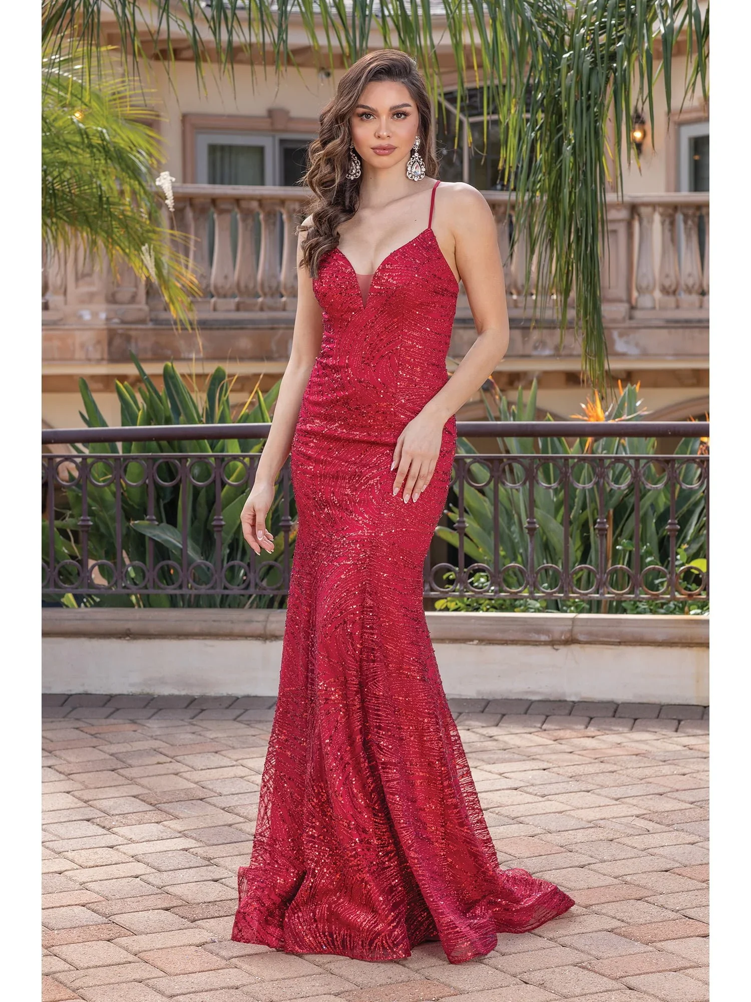 Woman in a red sequined evening gown standing on brick pavement with a tropical background of plants and a balcony.