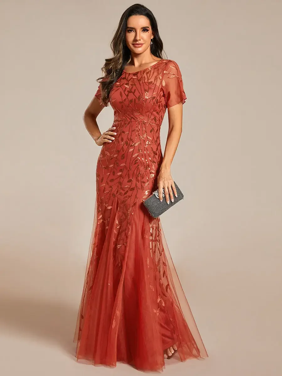 A woman wearing a long, elegant red dress with sheer fabric and gold leaf patterns, holding a small black clutch, standing against a neutral background.