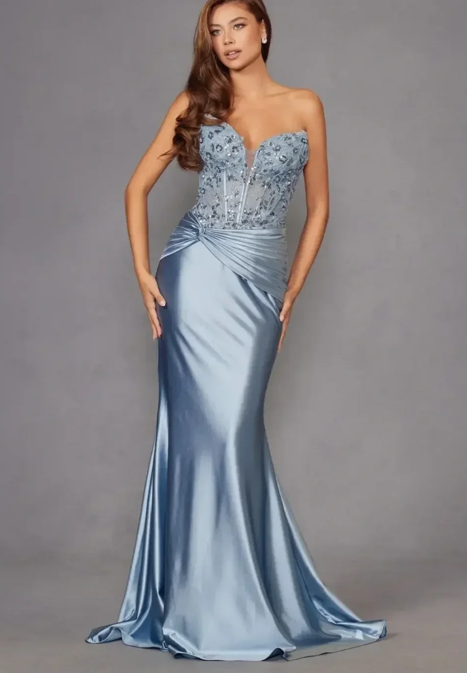 A woman in a strapless, light blue satin evening gown with floral embellishments on the bodice, standing against a plain gray background.