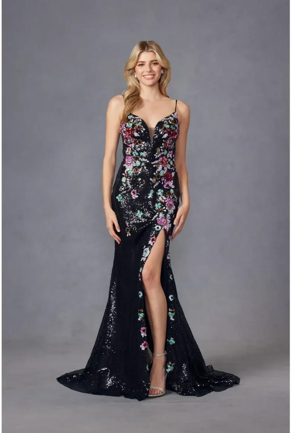 A young woman with long blonde hair wearing a black, floral, sequined evening gown with a thigh-high slit and spaghetti straps, standing against a gray background.