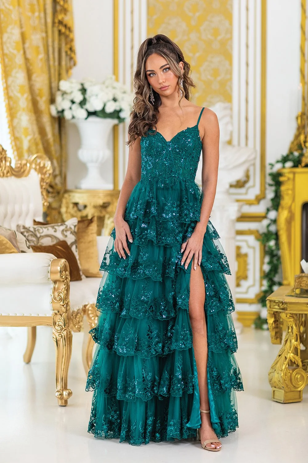 A woman in a teal, multi-tiered lace gown with a thigh-high slit, standing in a luxurious, gold-accented room with ornate furniture and floral arrangements.