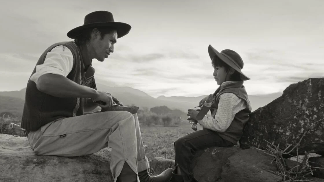 A man and a young boy sit outdoors, facing each other. The man, wearing a hat and vest, appears to be talking to the boy, who wears a hat, scarf, and jacket. The background features distant mountains and a cloudy sky.