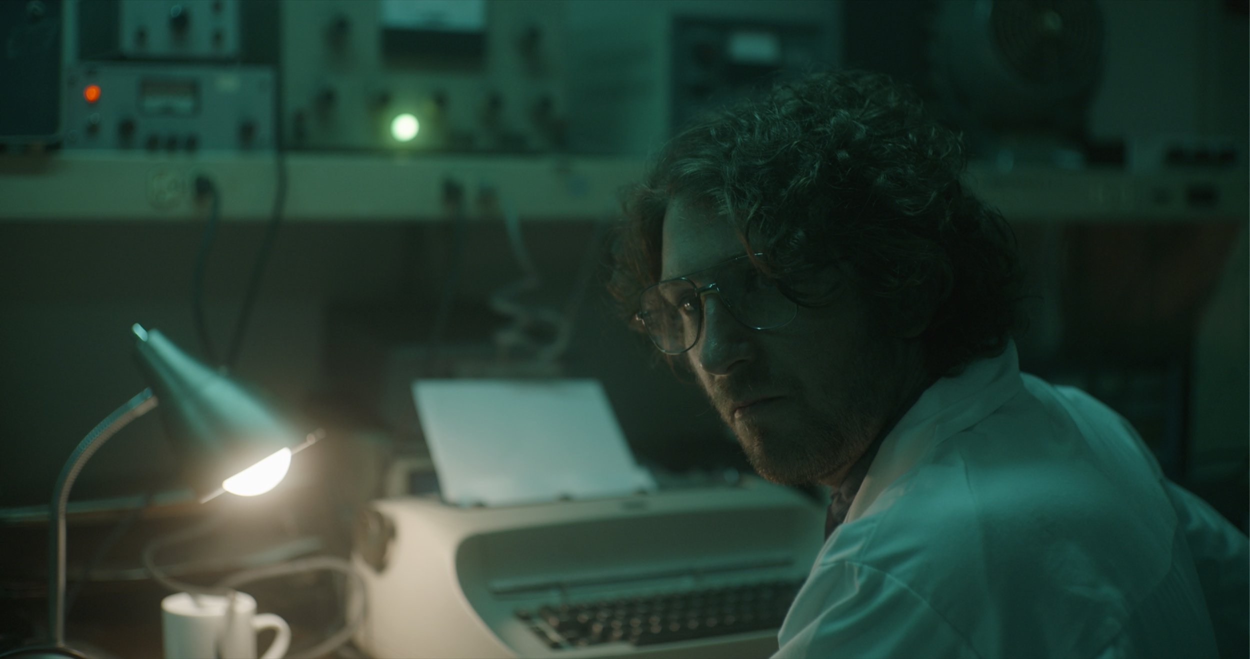 A man with curly hair and glasses working at a desk with a laptop, illuminated by a desk lamp, in a dimly lit laboratory or technical workspace.