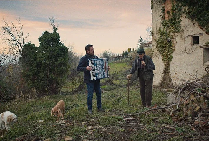 Two men, one playing an accordion and the other holding a walking stick, stand outdoors with a dog and a small goat nearby, in a rural setting with an old building covered in vines.