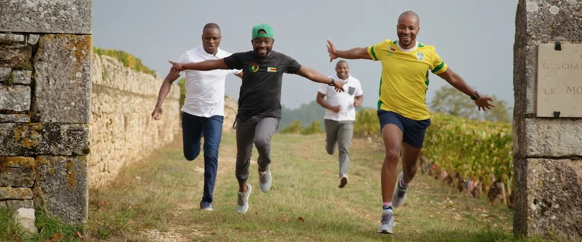 Four men running and smiling outdoors near a stone archway in a vineyard.