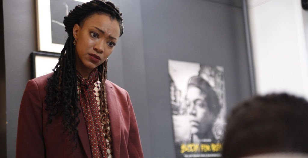 A woman with braided hair and a pink blazer looks concerned while talking to someone off-camera in a room decorated with black and white portraits.