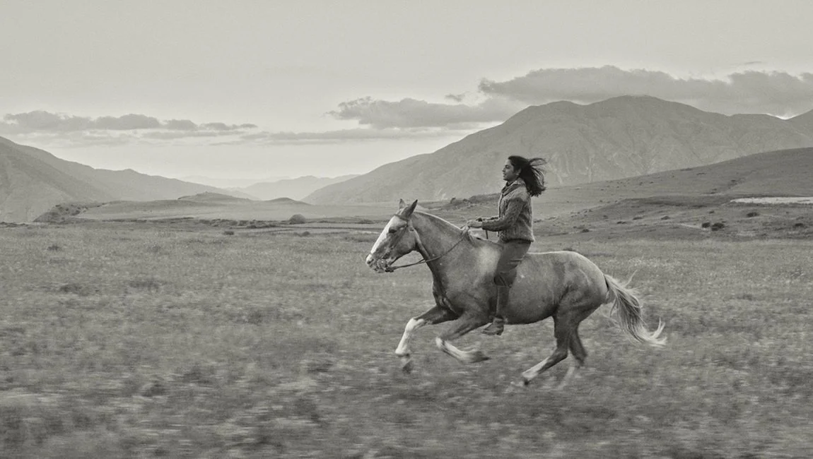 A woman riding a horse across an open field with mountains in the background, in black and white.