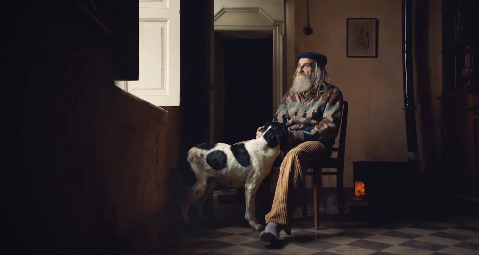 An elderly man with a gray beard sitting on a wooden chair, wearing a hat and patterned sweater, with a black and white dog standing beside him inside a dimly lit room near an open window.