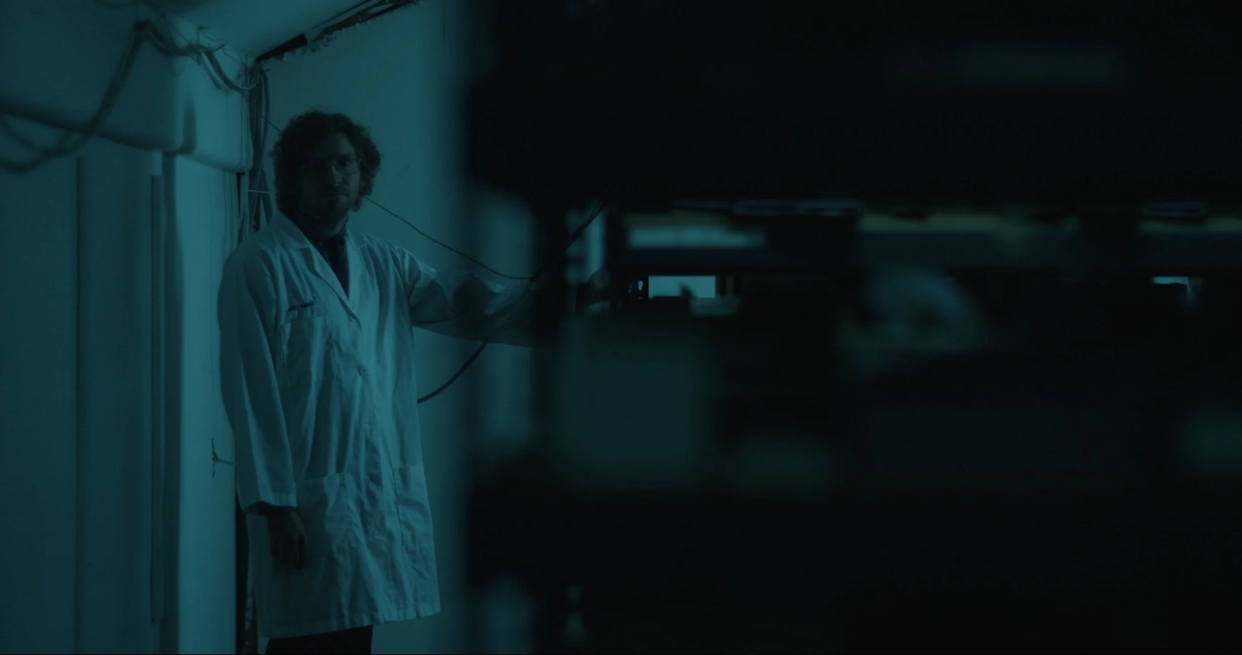 A man wearing a white lab coat and glasses standing in a dimly lit room, possibly a laboratory or research facility.