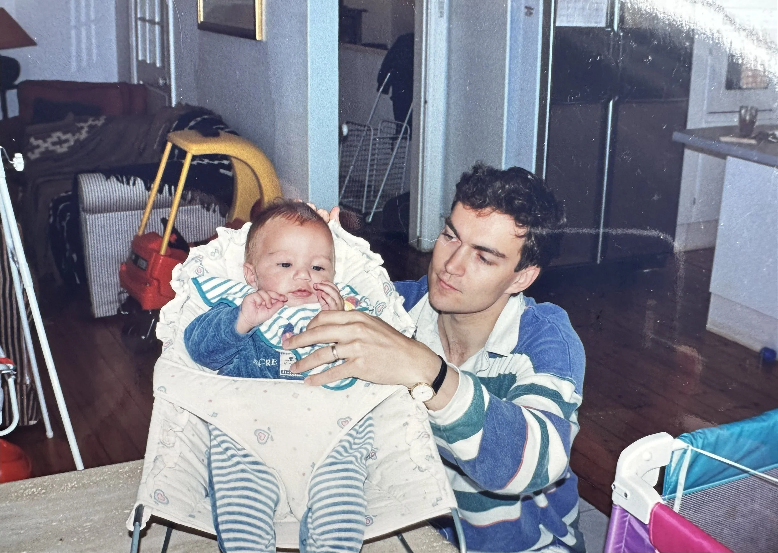 A young man with dark hair, wearing a blue and white striped shirt, holding a baby in a high chair indoors. The baby has light hair, is dressed in a denim jacket and striped bib, and is touching their face. The setting appears to be a kitchen or dining area with wooden flooring, a table surface in the foreground, and various household items and appliances in the background.