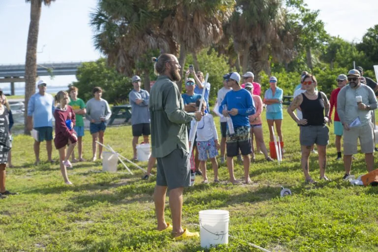 A large group of people gathered outdoors around a man speaking and holding a fishing rod, with palm trees and a bridge in the background.