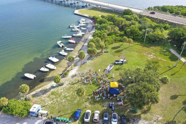 Aerial view of a shoreline park with boats docked along the water, people gathered near tents and picnic areas, and a highway in the background.
