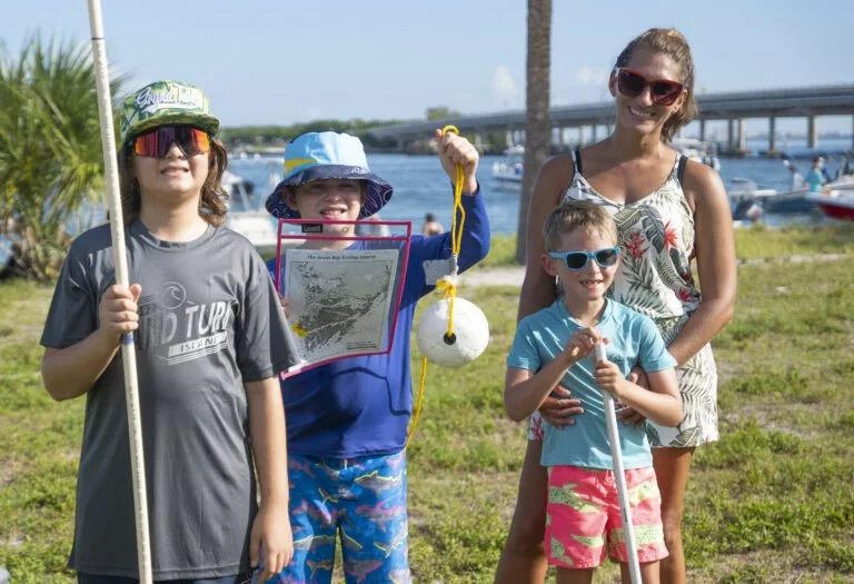 A woman with three children on a grassy area near water, holding fishing gear and a map, with boats and a bridge in the background on a sunny day.