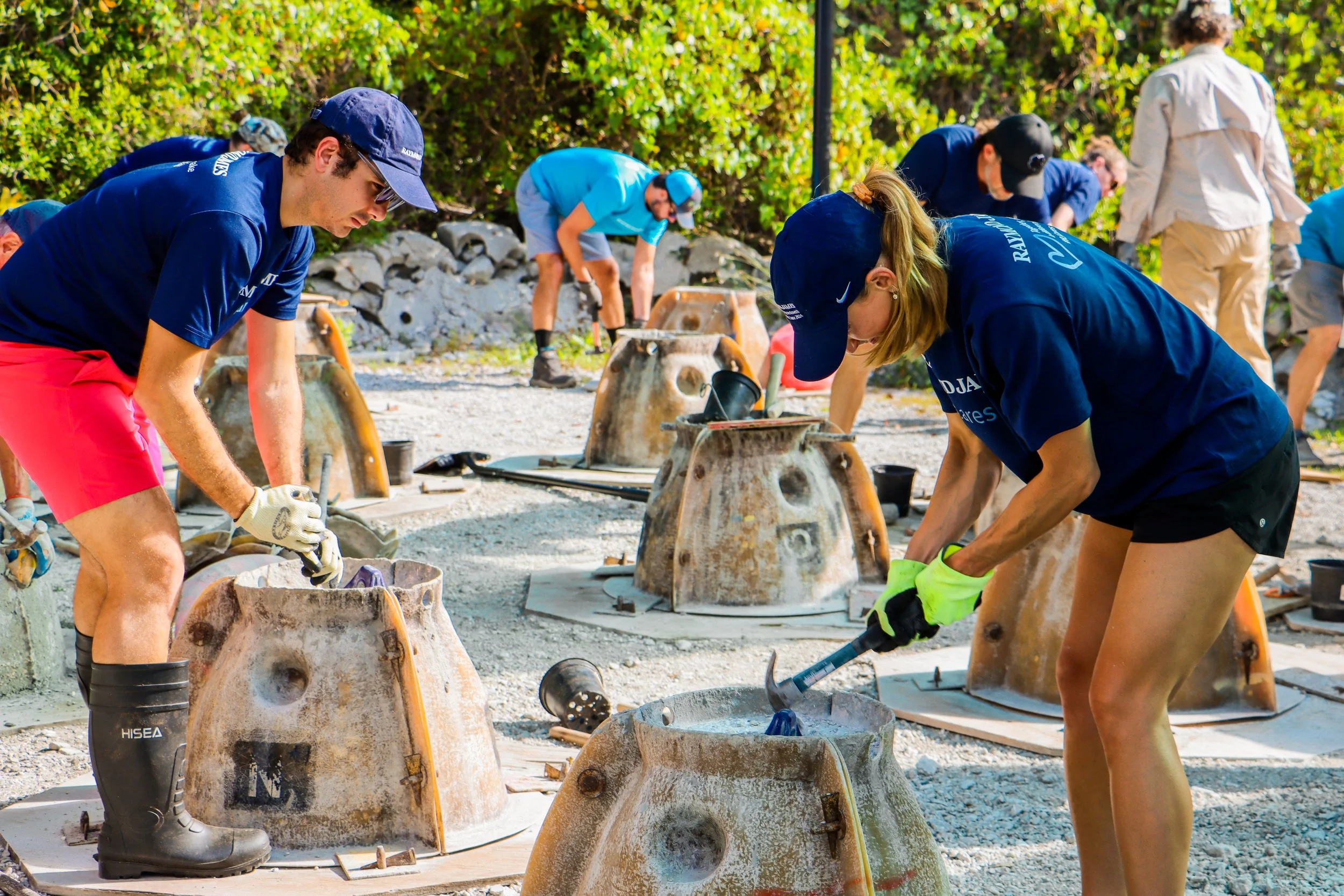 People working outdoors on a construction or cleanup project, wearing casual clothing and safety gear, surrounded by tools and equipment.