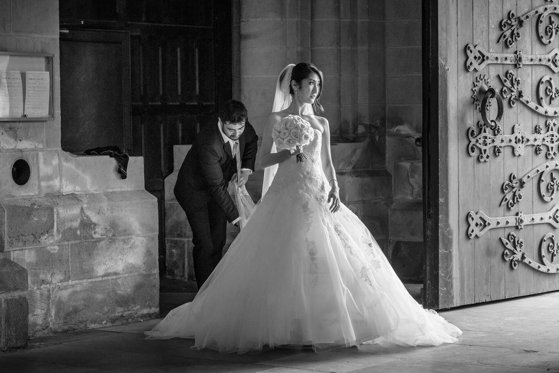 Une femme en robe de mariée tient un bouquet de roses, un homme l'aide à ajuster sa robe près d'une porte en bois ornée, dans une église.
