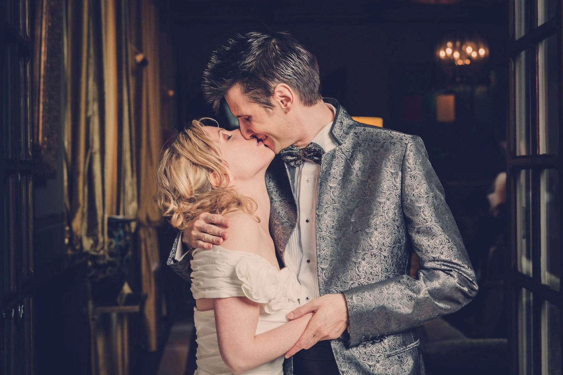 Un couple en costume élégant partage un baiser dans un intérieur luxueux.