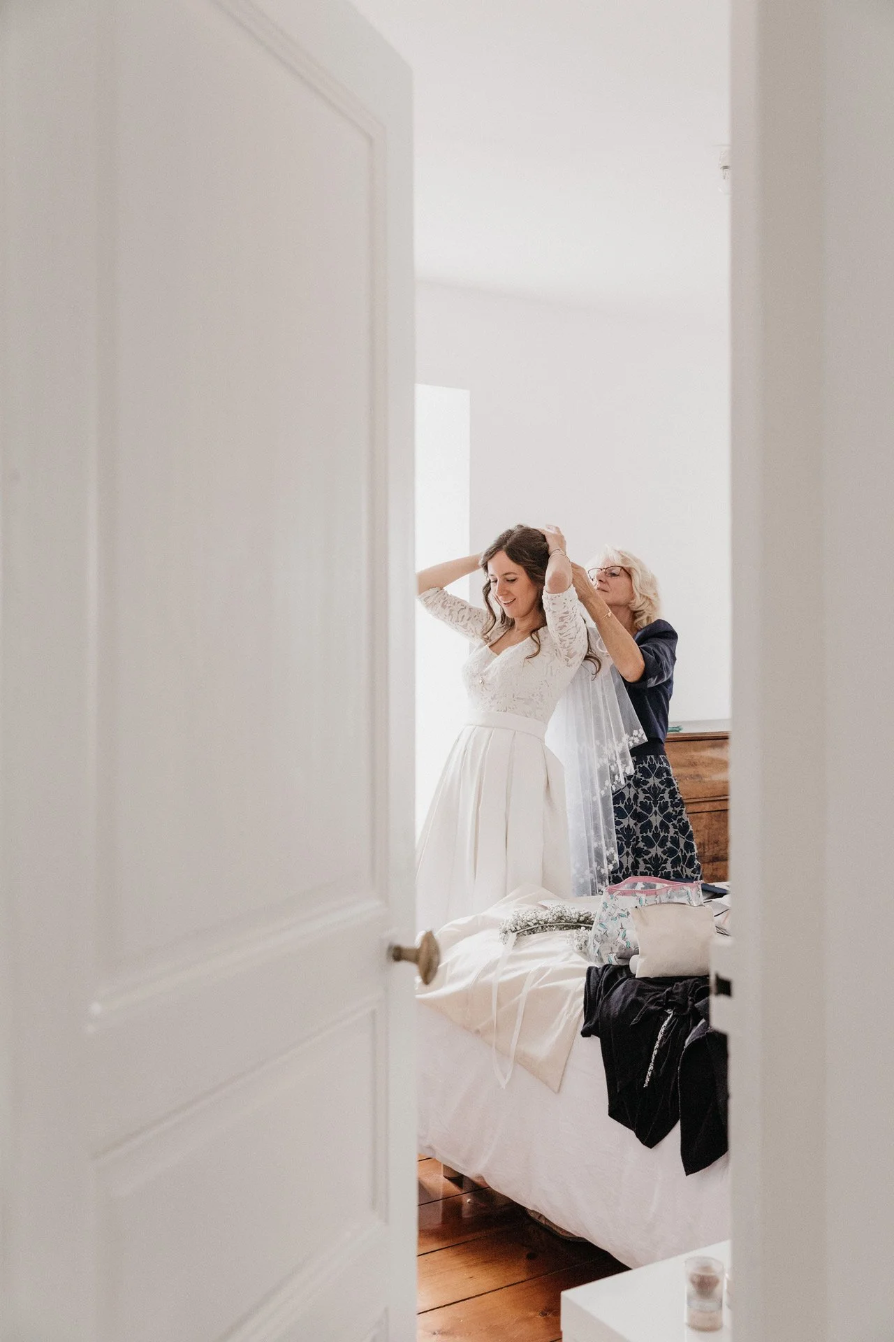 Une jeune femme en robe de mariée se prépare dans une chambre lumineuse, vue partielle à travers la porte entrouverte.