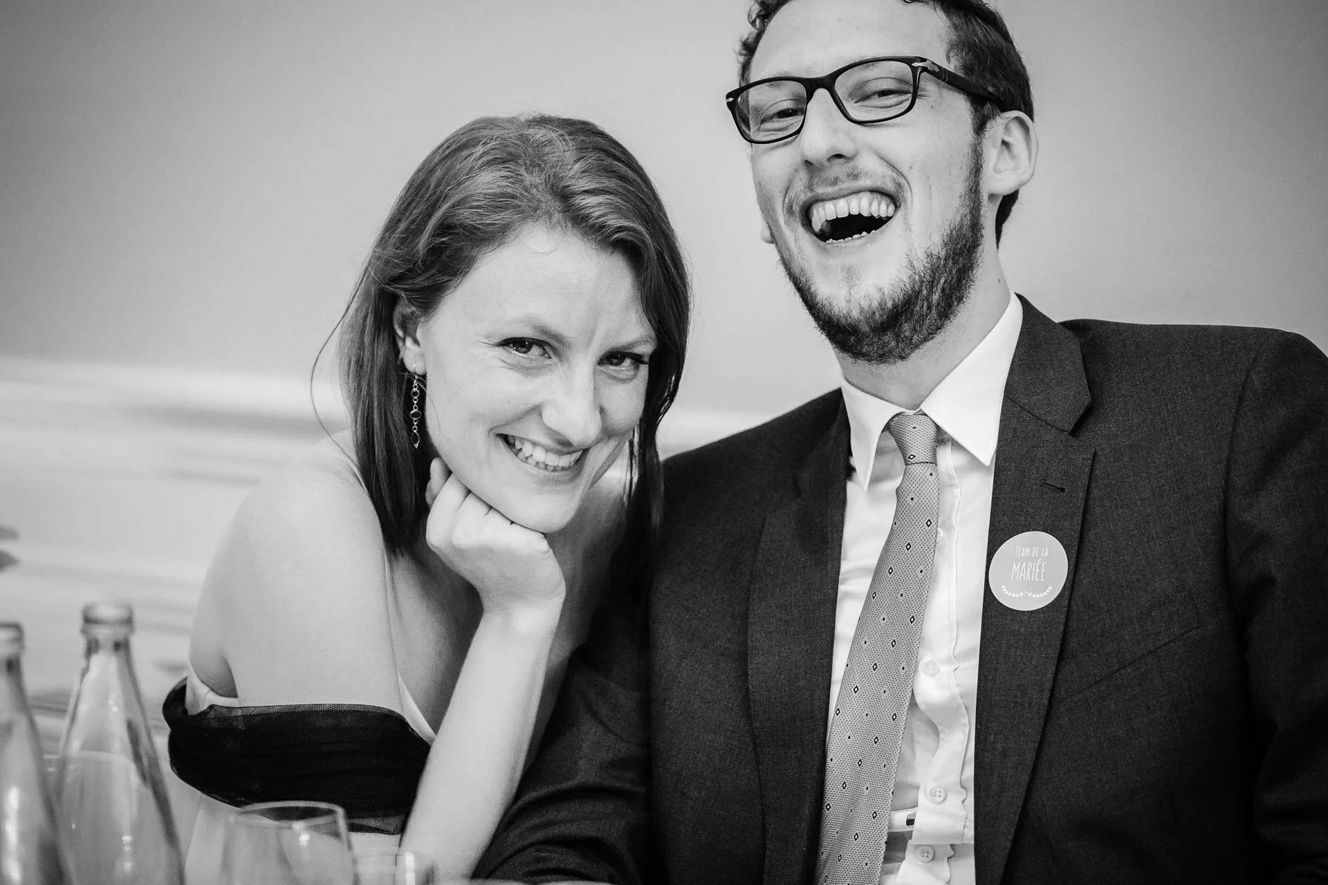 Un couple souriant, assis ensemble, lors d'un diner de mariage, en noir et blanc.