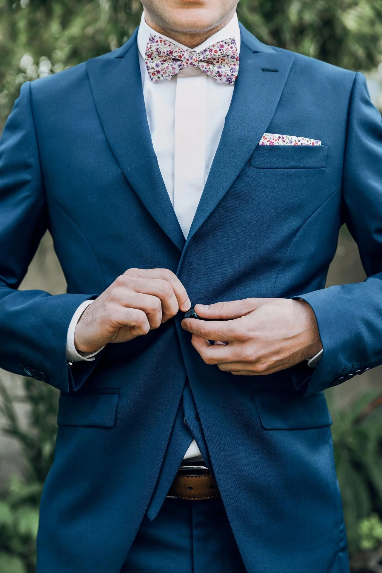 Futur marié en costume bleu ajustant la boutonnière, portant une chemise blanche, un nœud papillon à motifs floraux, une pochette de costume, et un pantalon en costume, dans un décor extérieur.