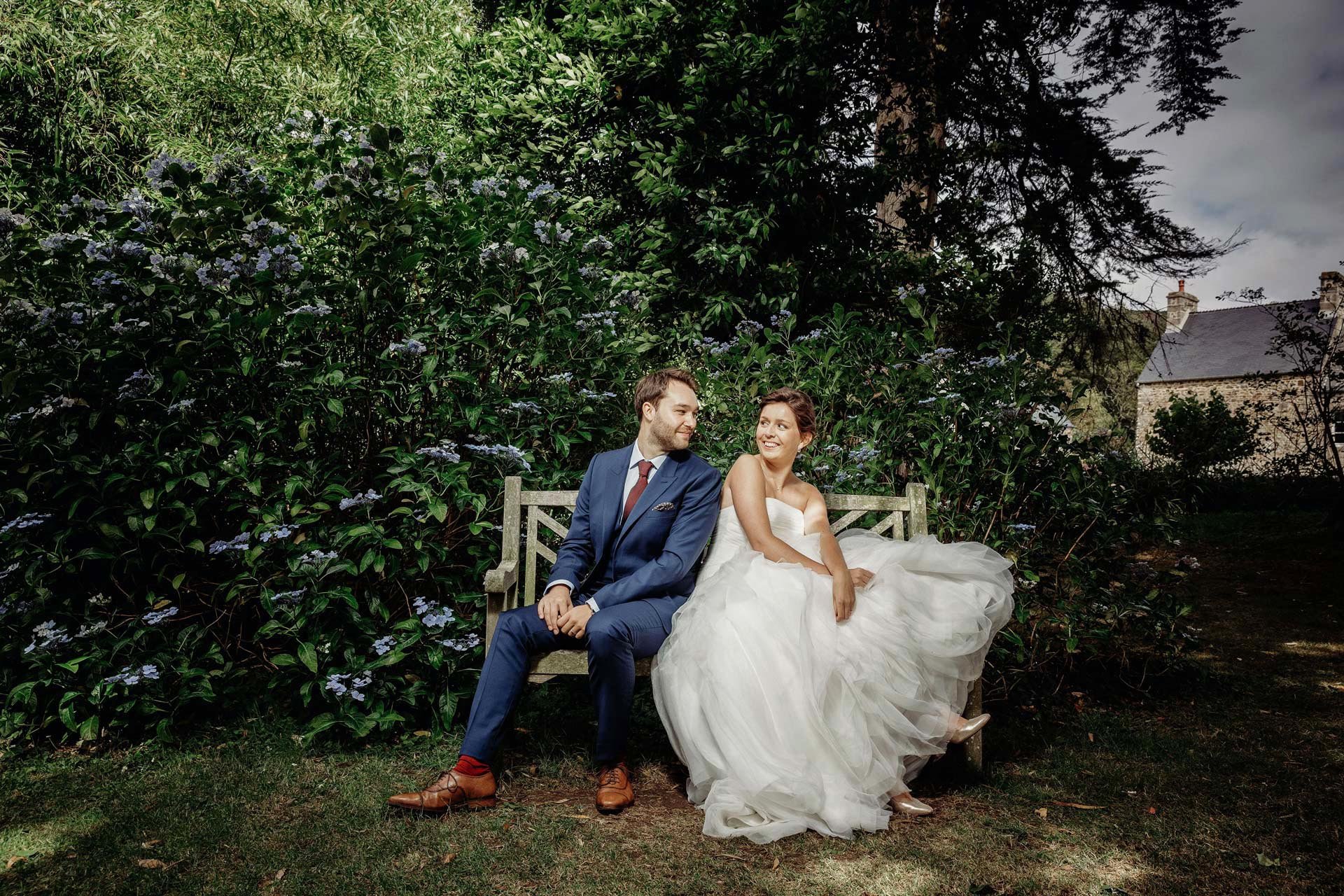 Un couple en habits de mariage assis sur un banc en plein air, entouré de végétation luxuriante, avec une maison en pierre en arrière-plan.